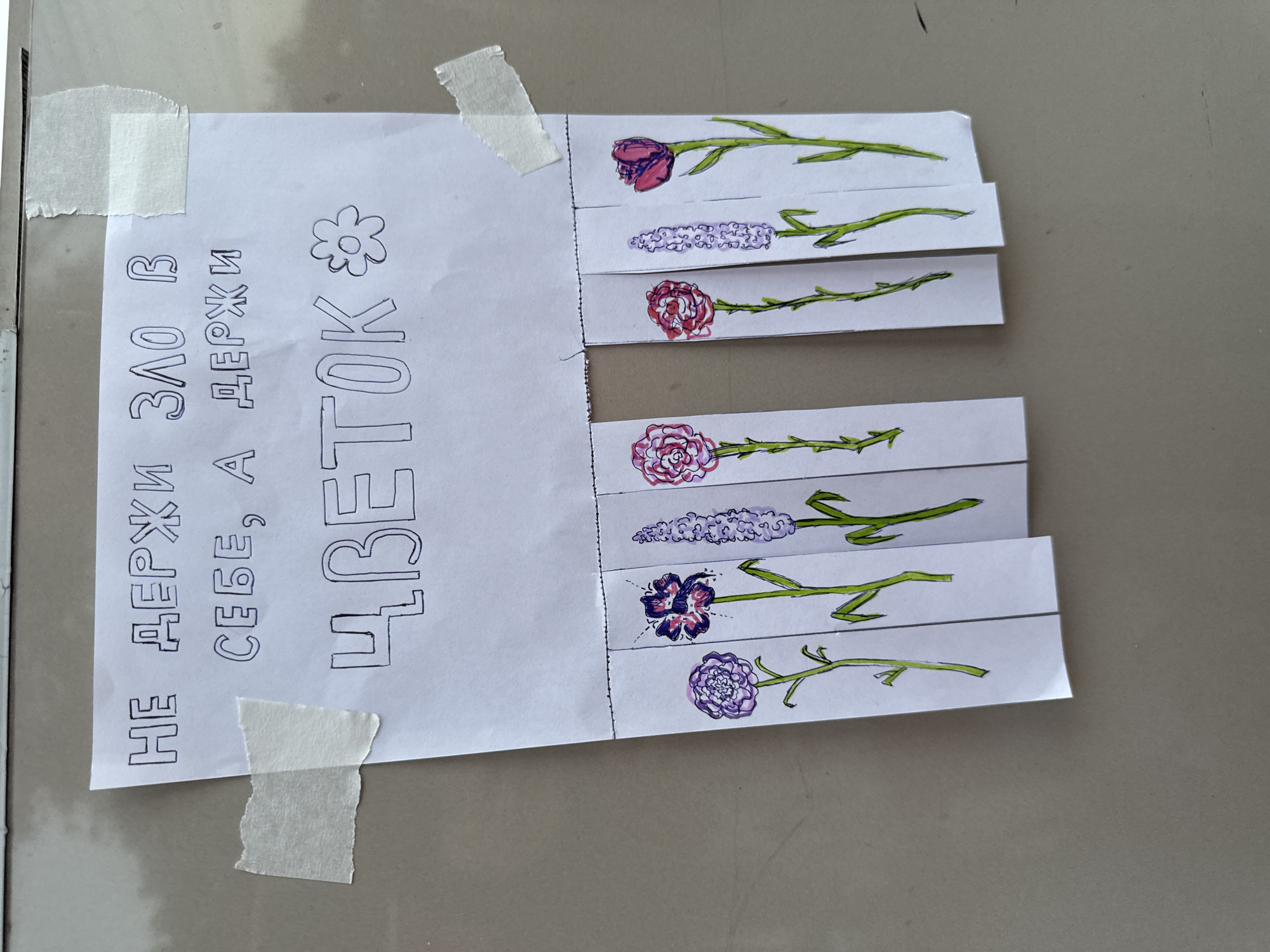 The image shows a piece of paper with a handwritten message and drawings, taped to a textured, possibly metallic, surface. The paper is secured with small strips of light-colored tape. The message, written in block letters, says "НЕ ДЕРЖИ ЗЛО В СЕБЕ, А ДЕРЖИ ЦВЕТОК" which translates to "Don't hold anger in yourself, but hold a flower." Below the message, there are tear-off strips, each featuring a drawing of a flower. The flowers are colored with purple, pink, and red hues, and have green stems. The overall presentation suggests a piece of street art or a whimsical flyer intended to be taken. The background surface appears to be a neutral grey, possibly a metal panel on a building or street furniture. There is no indication of the specific location within Sochi, Russia, or any people present in the image.