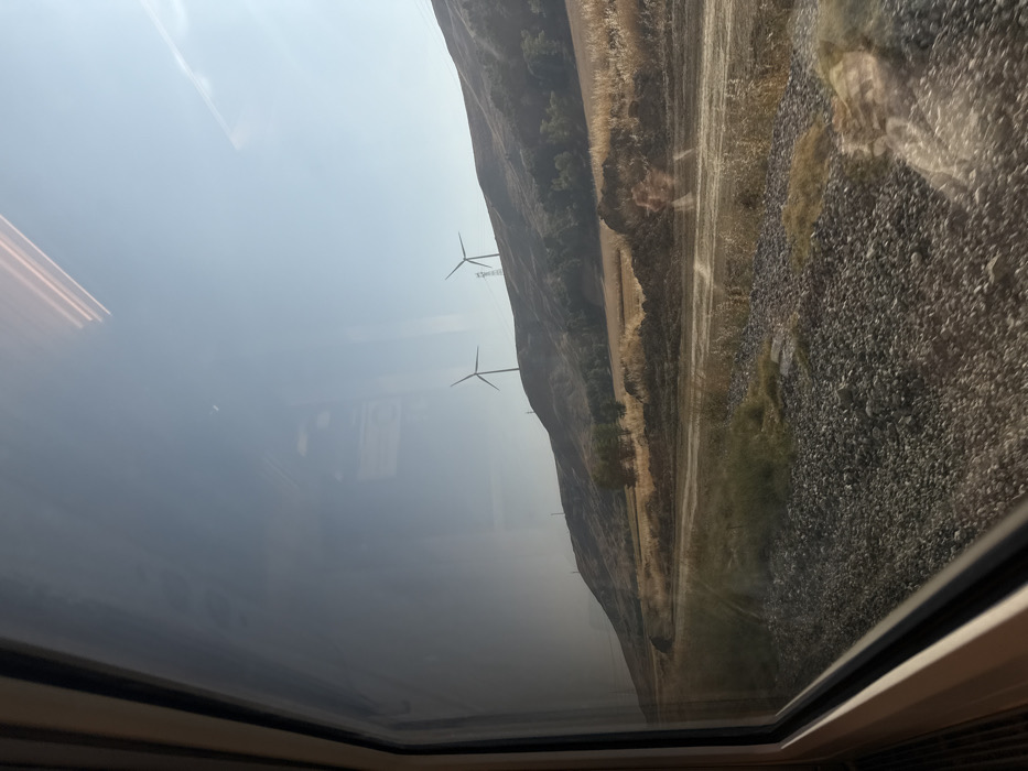 The image presents a view from inside a vehicle, likely a train, looking out a window at a rural landscape under a clear sky within the region of Palencia, Spain. The left portion of the frame is occupied by the vehicle's light-colored window frame, with reflections visible on the glass surface. Outside the window, the immediate foreground consists of a gravel-covered area, consistent with a railway track bed, running parallel to a dirt path or service road. Beyond this path, dry, yellowish-brown vegetation and scattered green foliage cover the terrain, with a mound of dark earth visible in the mid-ground. The background is characterized by undulating hills, extensively covered in parched, golden-brown grasses and intermittent patches of trees. Two large, white wind turbines are prominently positioned on separate hillcrests, and several utility poles are also visible along the ridges. The sky above is clear and bright blue, indicative of daylight. No individuals, animals, or explicit actions are discernible within the scene.