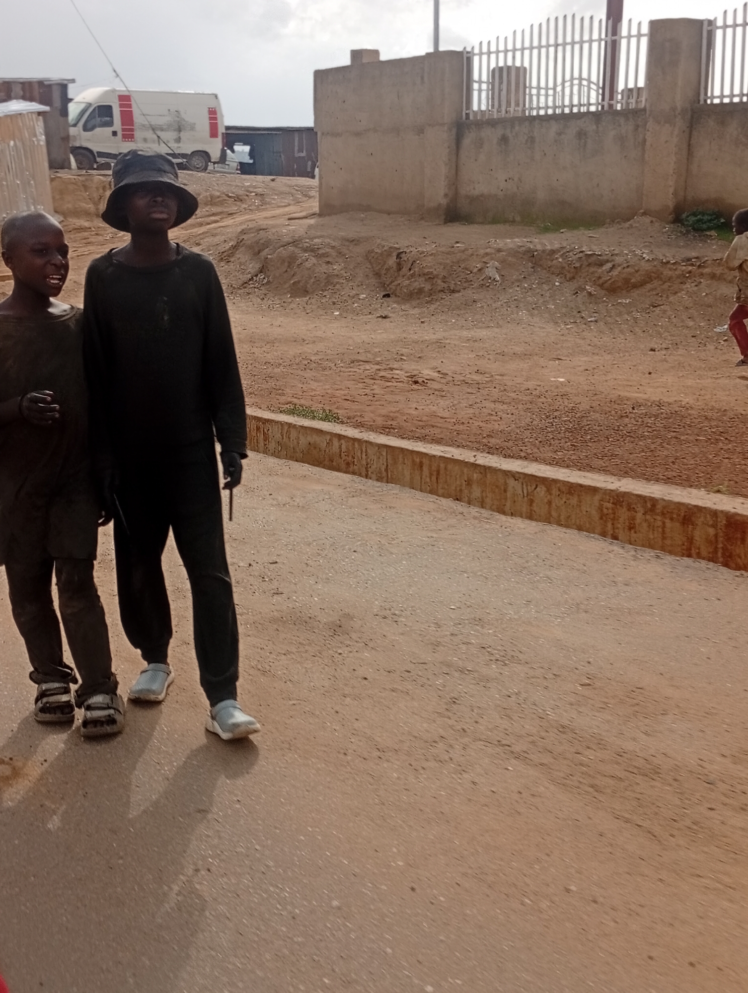 The image shows two young boys walking on a dirt road in Jos, Nigeria. The boy on the left is wearing a dark t-shirt and shorts, and sandals. The boy on the right is wearing a black long-sleeved shirt, black pants, and sneakers. He is also wearing a dark bucket hat. The boys appear to be walking away from the camera, and their expressions are not clearly visible.
The scene is an outdoor setting with a dirt road. In the background, there are some buildings made of corrugated metal and concrete. A white van with red stripes is parked in the distance. There is also a concrete wall with a metal fence on top. The ground is dry and dusty, suggesting a dry season or arid climate. The sky is overcast, indicating a cloudy day. The time of day appears to be daytime.
There is no visible text in the image. The overall impression is a candid shot of children in a seemingly impoverished or developing area. The focus is on the two boys, and their presence suggests a daily activity like walking to school, playing, or running errands.