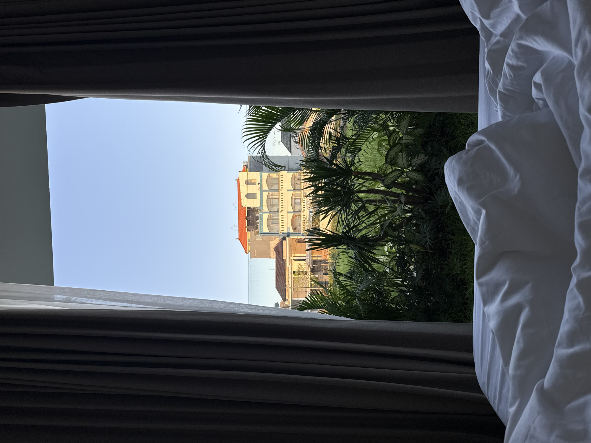 The image depicts a view from a bed through a window partially obstructed by dark gray curtains and white linens. Outside the window, several buildings with tan, orange, and white facades are visible, along with lush green vegetation including palm trees. The sky is a clear blue. Based on the architectural style and tropical vegetation, this scene is likely located in North Kuta, Indonesia. No people are visible.