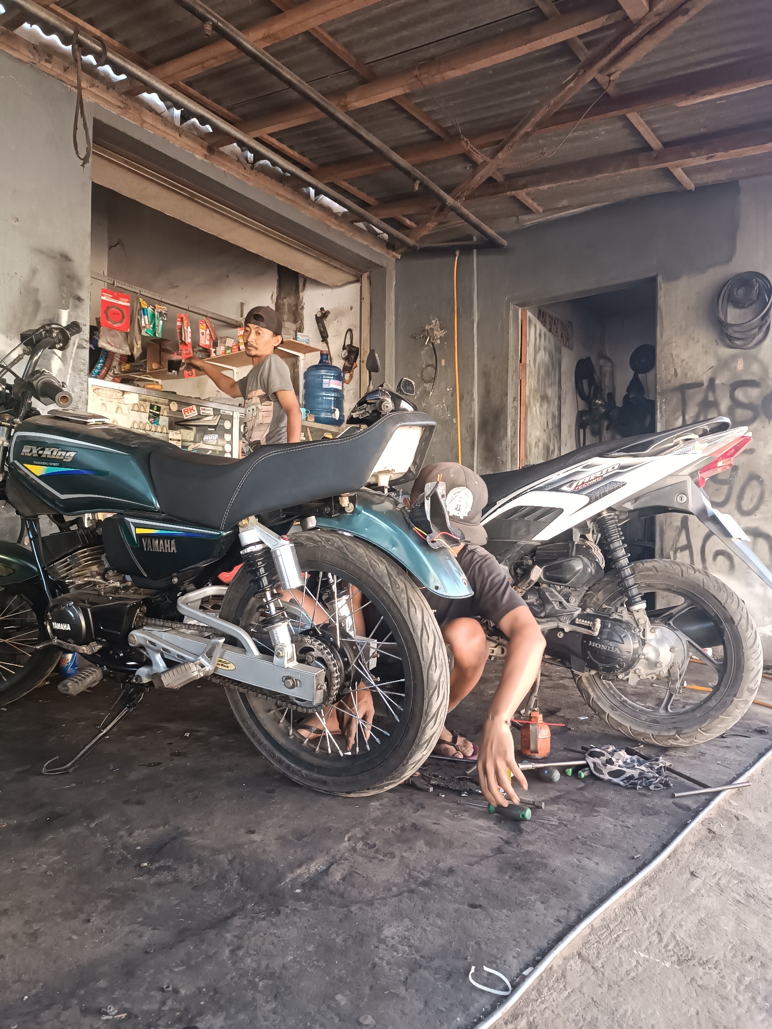 A full shot shows two men working on motorcycles inside a garage in Brongkal, Indonesia. The garage has a corrugated metal roof supported by wooden beams and concrete walls. The floor is made of concrete and is covered in grease and debris. A green Yamaha motorcycle is in the foreground on the left, and a white Honda motorcycle is in the background on the right. One man is kneeling beside the Honda, appearing to be working on its rear wheel, while the other man stands by the Yamaha, looking towards a shelf of parts. Both men are wearing casual clothing, and the kneeling man has a cap on his head. Tools and parts are scattered on the floor around the motorcycles.