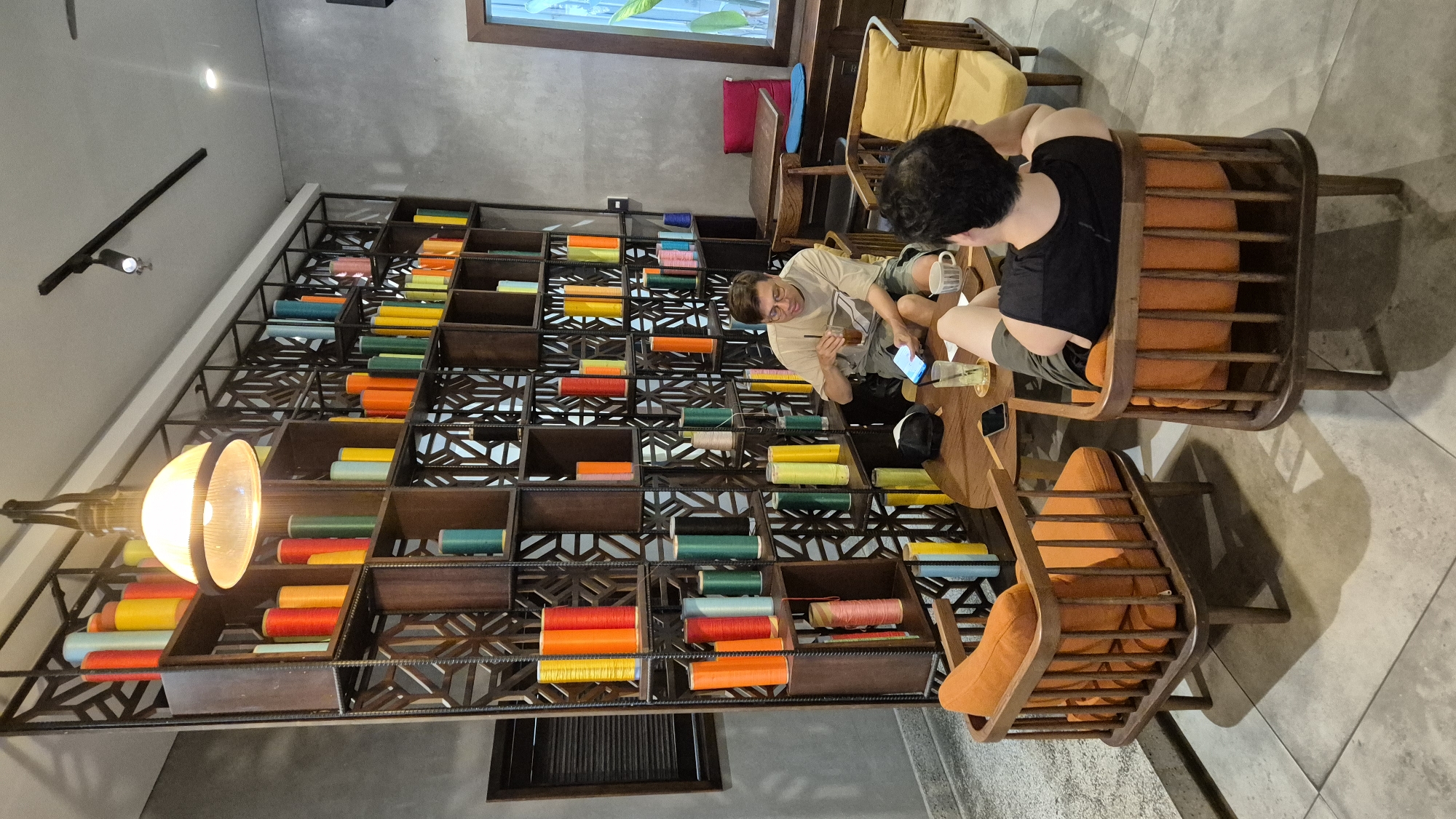 This image depicts two individuals seated in a cafe in Da Nang, Vietnam, with a prominent wall unit filled with colorful spools of thread. The wall unit, constructed of dark wood with a geometric lattice design, serves as a visually striking backdrop. Spools of thread in various vibrant colors, including red, orange, yellow, green, blue, and pink, are neatly arranged in the cubbies of the unit.
The cafe environment features concrete walls and tiled flooring. Two individuals are seated at a round wooden table. One individual, a man wearing glasses and a light-colored t-shirt and shorts, is holding a drink with a straw and looking at a smartphone. The second individual, whose back is to the camera, is wearing a black sleeveless shirt and shorts and is seated in an orange-upholstered wooden chair. Other seating in the cafe includes an orange armchair and a yellow armchair. A wooden cabinet with a red cushion and a blue cushion is visible in the background.