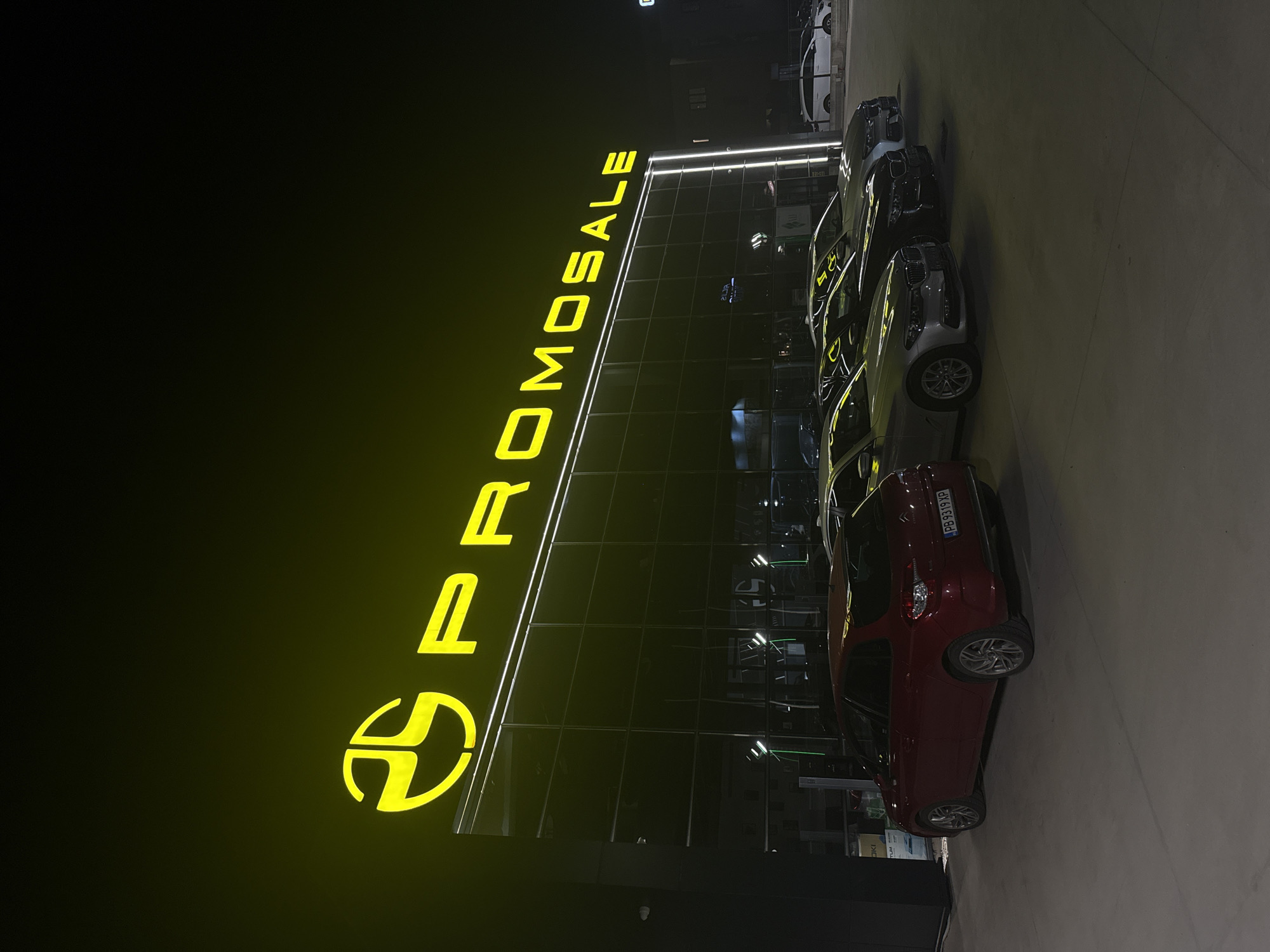 The image shows a nighttime view of the "PROMOSALE" dealership in Plovdiv, Bulgaria. The dealership's yellow illuminated sign is prominently displayed on the building. Several cars are parked in front of the dealership. The cars are various models and colors, including a red car in the foreground with license plate PB9319XP. The dealership building has glass walls. The surrounding area is dark, with streetlights providing some illumination.
