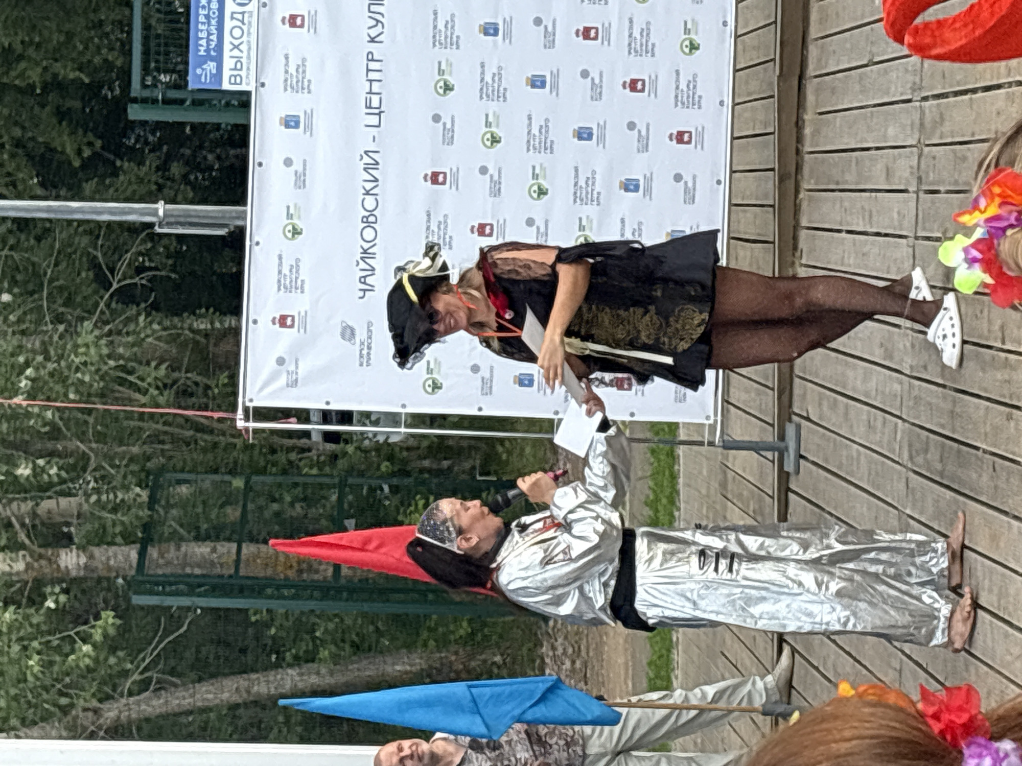 In an outdoor setting, likely at an event in Chaikovsky, Russia, two women are standing on a wooden stage. A large banner with repeating logos and text, including "ЧАЙКОВСКИЙ - ЦЕНТР КУЛЬТУРЫ" (Chaykovsky - Center of Culture), serves as the backdrop. To the left, a man in light-colored pants and a patterned shirt stands near a blue flag.
The woman on the left wears a shiny silver outfit, resembling a jumpsuit, and a white bandana. She is holding a microphone and a piece of paper, appearing to be speaking or presenting. She is barefoot.
The woman on the right is dressed in a black pirate-inspired costume, complete with a pirate hat and sunglasses. She wears black fishnet stockings and white crocs. She is also holding a piece of paper and looking towards the woman on the left, suggesting an interaction or handover.
The environment appears to be a park or open-air venue, with trees and foliage visible in the background, and what looks like a fence and poles. Several red and blue flags are also present. The overall impression is of a public gathering or festival.