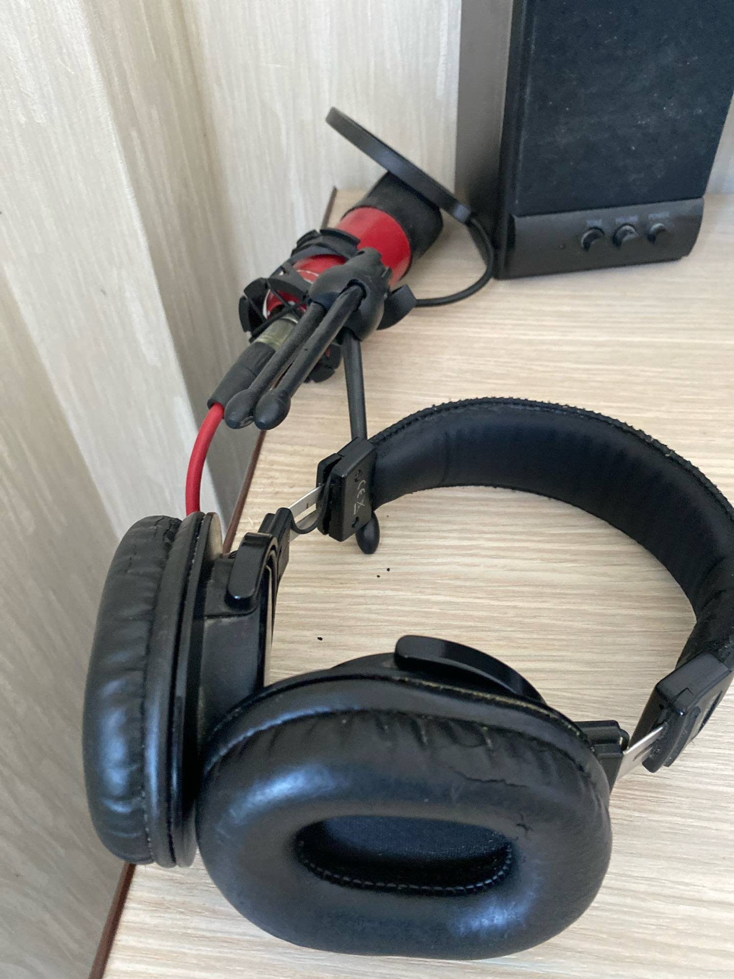 The image shows a pair of black over-ear headphones and a small red microphone with a black tripod stand, resting on a light-colored wooden surface. A black speaker is visible in the background to the right. The environment appears to be indoors, possibly a desk or shelf, with a textured white wall in the background on the left. There are no people or any indication of activity in the scene. The image does not provide any information about the city of Chișinău, Moldova.