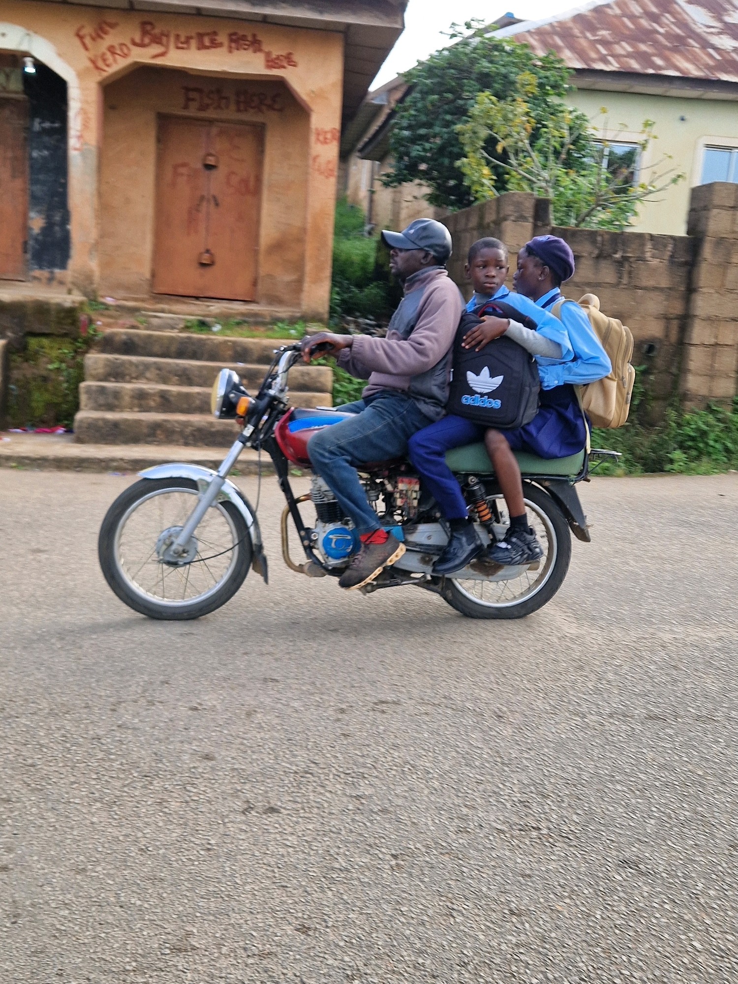 In Kabong, Nigeria, a man wearing a gray jacket and blue jeans rides a motorcycle. He is accompanied by two children, a boy and a girl, also on the motorcycle. The boy, in the middle, wears a black Adidas backpack. The girl, behind the boy, is in a blue school uniform and carries a tan backpack. They are traveling on a paved road, with buildings and trees visible in the background. The scene depicts a common mode of transportation for children and adults in the area.