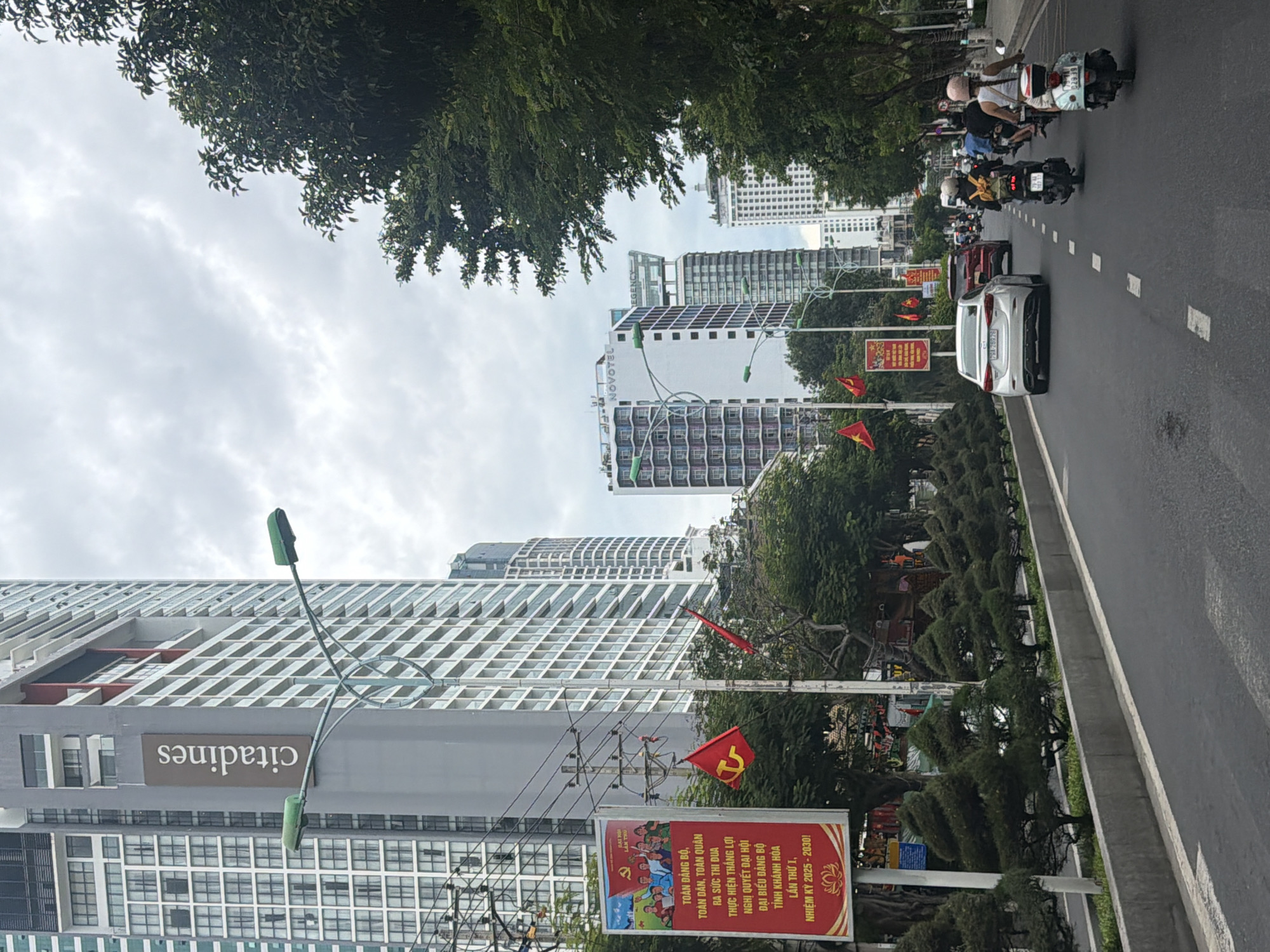 The image depicts a street scene in Nha Trang, Vietnam, characterized by modern architecture and bustling traffic. On the left side of the frame, a tall, grey building with numerous windows, identified by a "citadines" sign, dominates the skyline. Adjacent to it, a large red billboard features Vietnamese text and imagery, including a red flag with a yellow hammer and sickle, suggesting a political or cultural message. Several Vietnamese flags are also visible on poles.
The street itself is paved and marked with white dashed lines, indicating traffic lanes. A line of neatly trimmed, conical-shaped trees runs along the side of the road, separating it from a concrete barrier. In the foreground, several vehicles are in motion: a white sedan, a red SUV, and multiple motorcycles. The motorcyclists are diverse in their attire, with some wearing helmets and backpacks. The sky is overcast, indicating a cloudy day.
Further down the street, more high-rise buildings are visible, including one with "NOVOTEL" signage and another with "REGENCY" labeling, pointing to a commercial and tourist-oriented area within Nha Trang. The presence of numerous modern buildings, traffic, and flags suggests a developed urban environment.