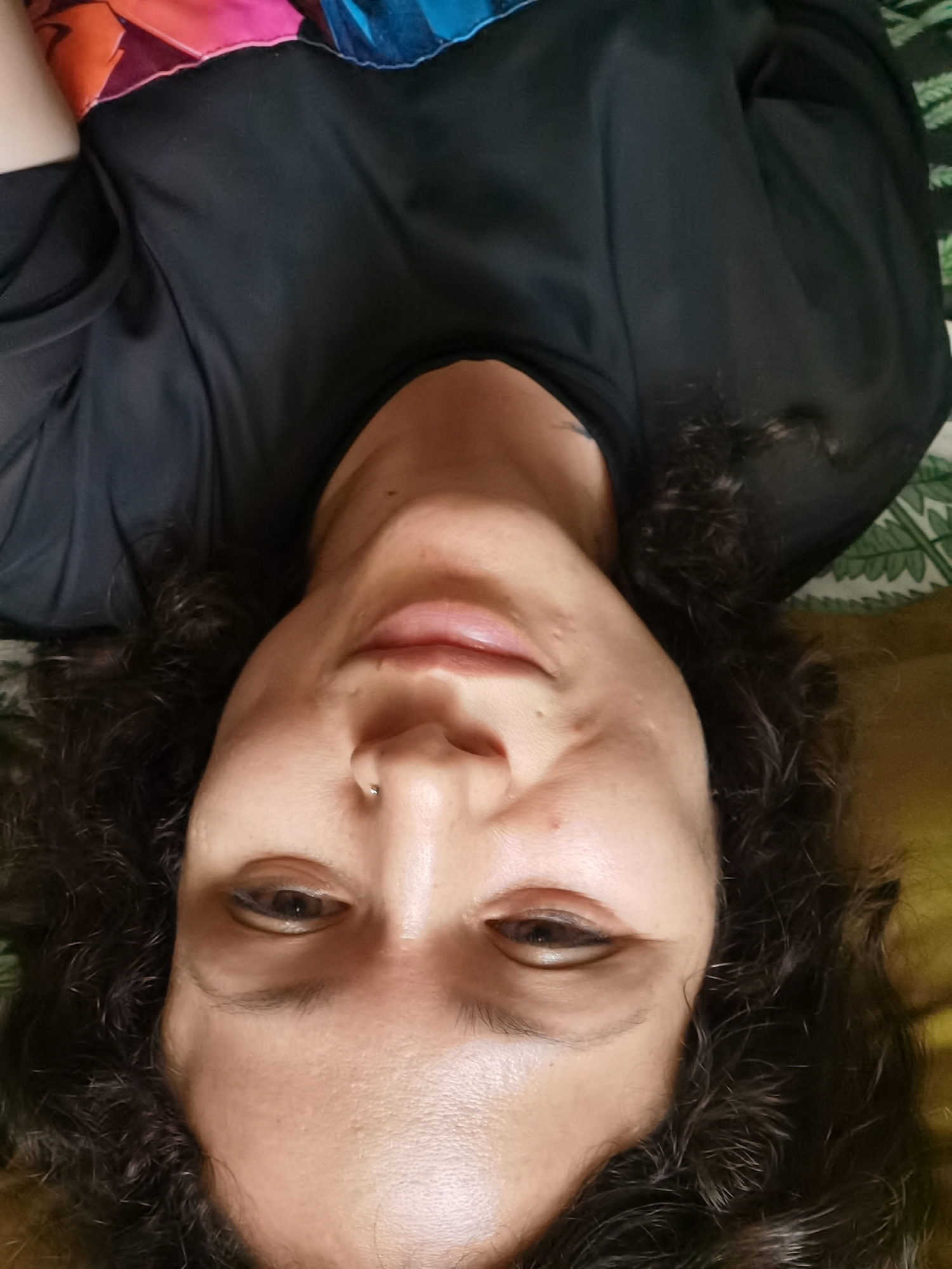 The image shows a close-up, upside-down selfie of a person with curly, dark hair. The person is wearing a black top with a colorful, patterned neckline. Their face is partially visible, with their eyes, nose, and mouth in focus. The background appears to be a bed with a patterned sheet and a yellow pillow. The lighting is soft and diffused. There is no discernible urban environment or specific location within Chaikovsky, Russia.