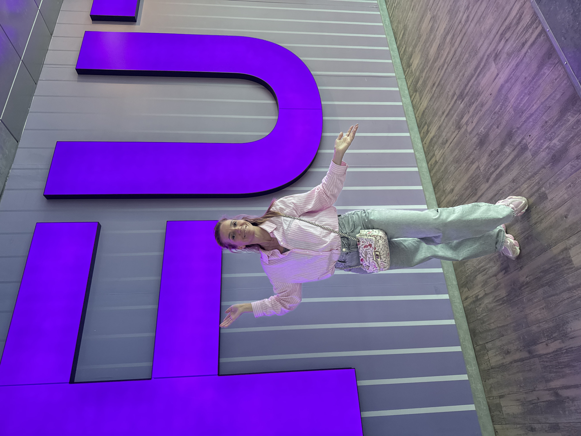 A medium shot depicts a woman standing in front of a large, purple architectural display. The display appears to be a modern art installation featuring oversized letters. The woman is wearing a pink striped shirt, light-wash jeans, and pink sneakers. She has a small patterned purse slung across her body. She is smiling and has both arms raised with open palms facing forward. The background is a wall with horizontal grey and white stripes, suggesting an interior space. The floor to the right is wooden. The scene suggests a photo opportunity or an interactive art exhibit. Based on the architectural style and color scheme, it is plausible that the location is within a modern exhibition or cultural center. Without further context or identifying marks, it is not possible to confirm that this photo was taken in Saint Petersburg, Russia.
