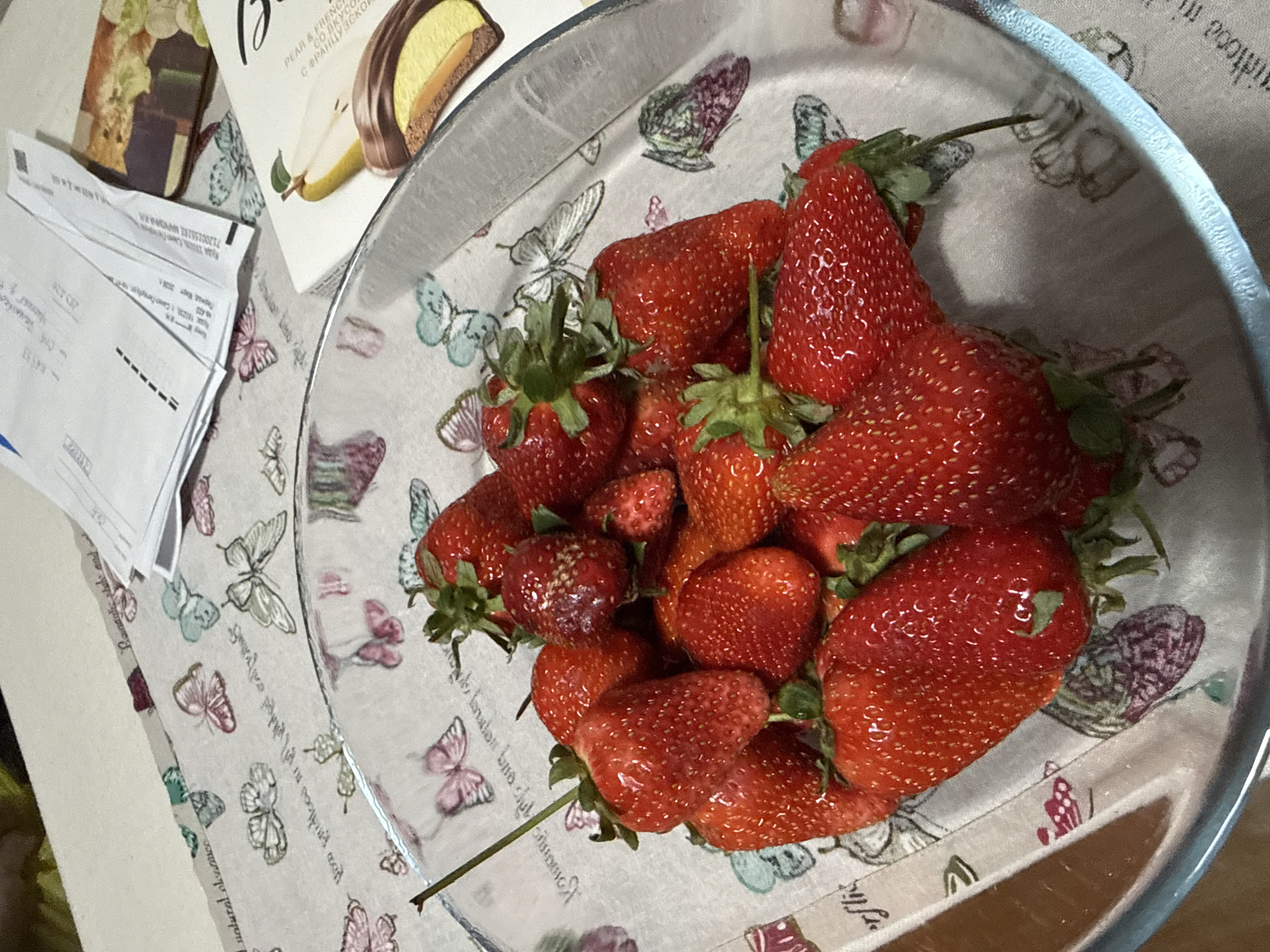 Here is a detailed description of the image:
The image depicts a bowl of fresh strawberries, a box of what appears to be pear and French chocolate candies, and some papers. The strawberries are various sizes and arranged in a glass bowl. The box of candies is partially visible, with the label "PEAR & FRENCH" printed on it. The papers appear to be documents or receipts, with some text visible, including the address "Saint Petersburg, Russia" written in Russian. The overall scene suggests a snack or food setting, possibly indoors. The tablecloth has a butterfly pattern.