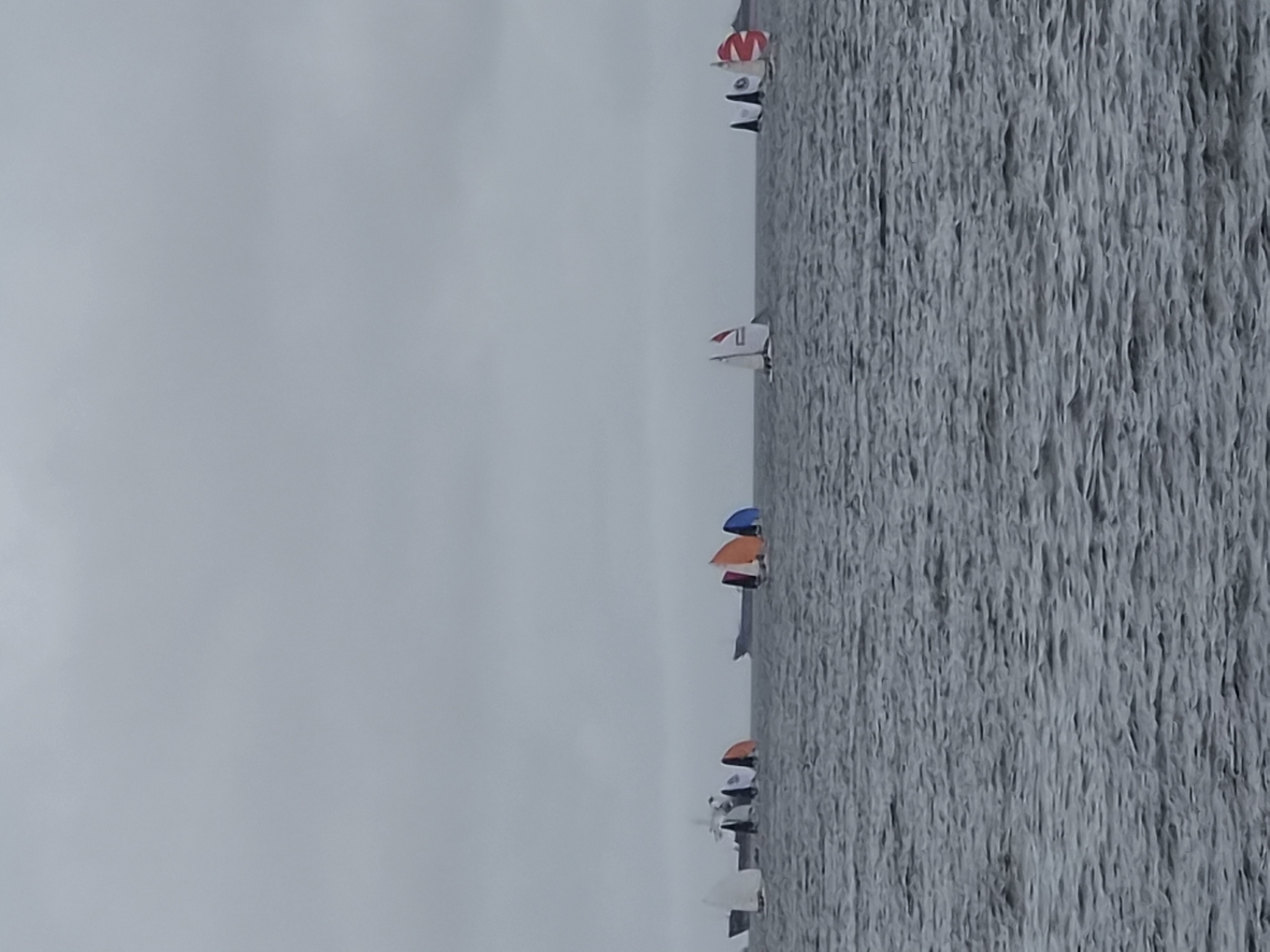 The image depicts a fleet of sailboats on the water, under an overcast sky. The sailboats have colorful sails, including orange, blue, and red. In the background, there are larger ships and a hazy shoreline, suggesting an urban or industrial setting. The water is choppy, indicating a moderate wind. The scene appears to be a regatta or a sailing event. The presence of larger vessels and the general atmosphere suggest this location might be a harbor or a bay. Without further information, it is not possible to pinpoint the exact location within Vladivostok, Russia, but the scene is consistent with its coastal geography and maritime activity.