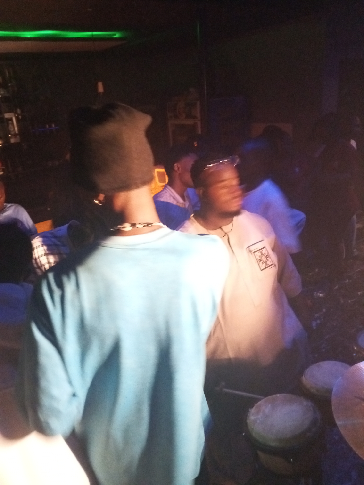 The image depicts a dimly lit interior, possibly a bar or nightclub, in Lokoja, Nigeria. Several people are present, with a man in a light blue shirt and dark beanie standing prominently in the foreground, facing away from the camera. Another man in a cream-colored shirt with a decorative square patch on the chest is visible to the right, his face blurred, suggesting movement. In the foreground, a set of drums is visible. The background is crowded with other individuals, some partially obscured. Above, a green light fixture illuminates the ceiling.
