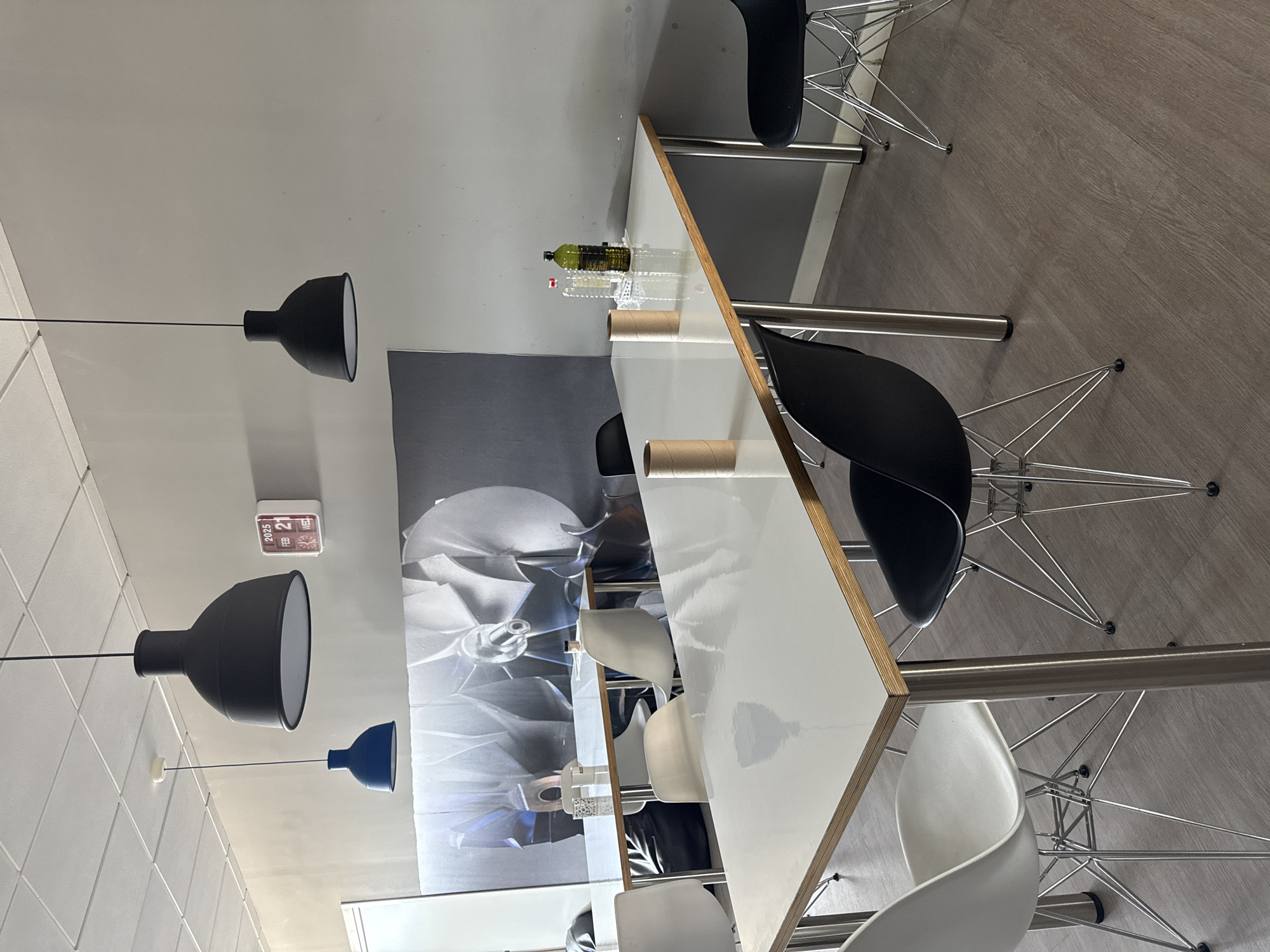 This image depicts a modern office or co-working space with a minimalist aesthetic, likely located in Getafe, Spain. The scene features two long, white-topped tables with chrome legs, arranged parallel to each other. Several chairs are placed around the tables, including white Eames-style chairs and black Eames-style chairs. The walls are painted a neutral gray, and a large, black and white mural of a turbine or fan is prominently displayed on the back wall. Three modern pendant lights hang from the ceiling, two large black ones and one smaller blue one. On one of the tables, there are two cardboard tubes, a clear plastic bottle, and a green glass bottle, suggesting recent use or preparation for an activity. A white digital clock displaying "2025 FEB 21 WED" is mounted on the wall. The flooring appears to be a light wood-look laminate. The space is well-lit, with a ceiling grid light system visible. No people are present in the image.