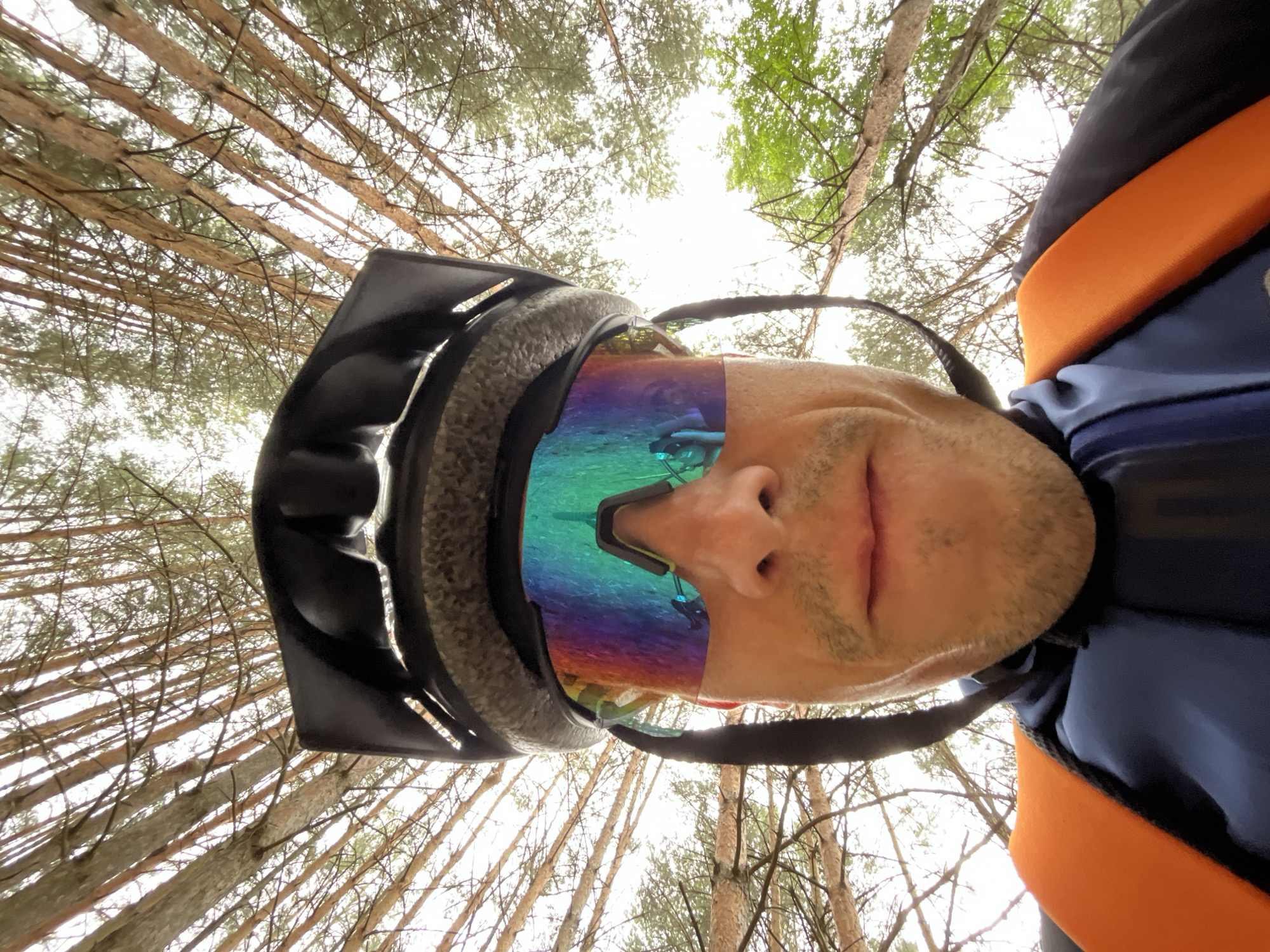 The image shows a first-person perspective of a person wearing a black bicycle helmet and brightly colored mirrored sunglasses. The person is looking upwards, with the focus on their face and the sky visible through the trees. The environment appears to be a forest with tall, thin trees and a bright, overcast sky. The person is wearing a dark blue jacket with orange straps from a backpack visible over their shoulders. No specific location in Chaikovsky, Russia can be determined from the image.