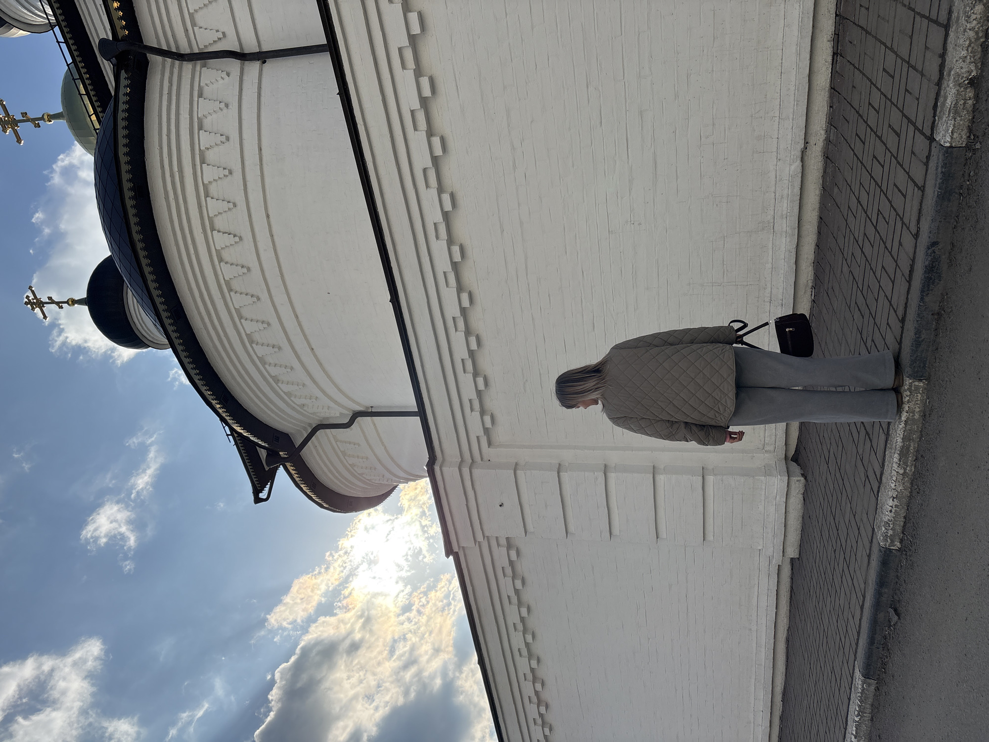 A low-angle, outdoor shot depicts a woman standing near a white building in Ulyanovsk, Russia. The woman, facing away from the camera, is wearing a light brown quilted jacket and light gray pants. She carries a black purse on her shoulder. The white building has a distinctive architectural style, with tiered layers and decorative elements. A dome with a cross is visible at the top of the building. The sky is partially cloudy, with the sun shining through. The woman is positioned on a gray tiled sidewalk next to a street.
