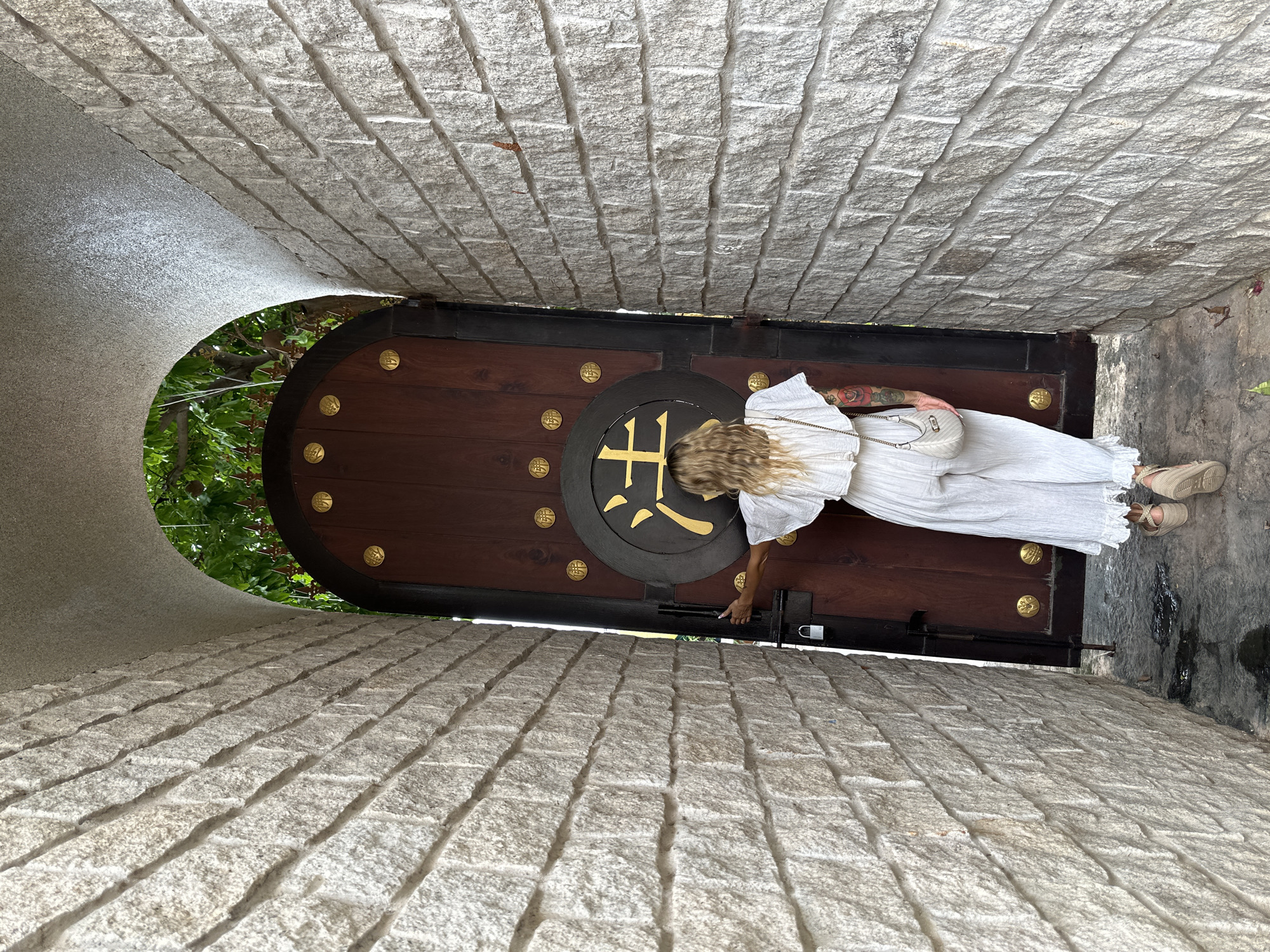A woman dressed in white stands in a narrow stone passageway in Mũi Né, Vietnam, in front of a large, arched wooden door adorned with a black circle featuring a golden, Japanese-style symbol. The door is also decorated with several smaller golden ornaments. The passageway walls are made of light-colored stone bricks arranged in a linear pattern. The woman is seen from the waist up, wearing a white dress and carrying a small white purse on a chain. Her hands are near a metal latch on the door, suggesting she is either opening or closing it. Green foliage is visible beyond the archway of the door.
