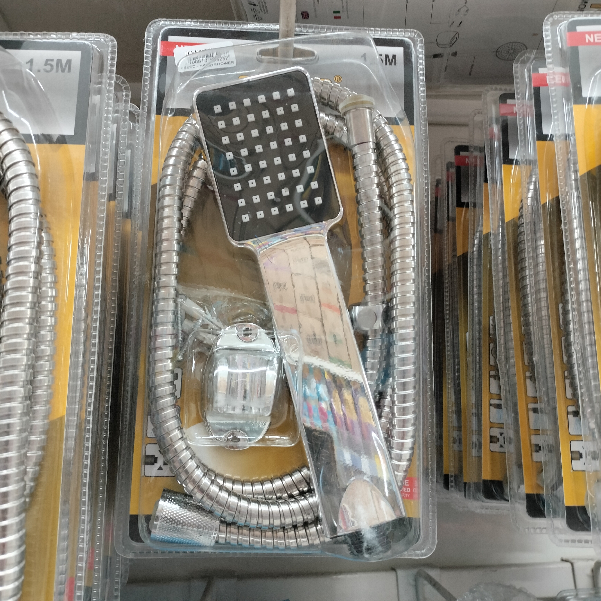 A retail display features multiple packaged shower head sets. Each set includes a chrome-finished hand shower head with multiple square nozzles, a flexible metal hose, and a wall-mounted holder, all contained within clear plastic packaging with yellow cardboard backing. Some packages show printed instructions and product information. The display appears to be in a store located in Tangerang, Indonesia, as indicated by subtle environmental cues and common retail product packaging styles found in the region. The lighting is bright and even, typical of indoor retail environments.