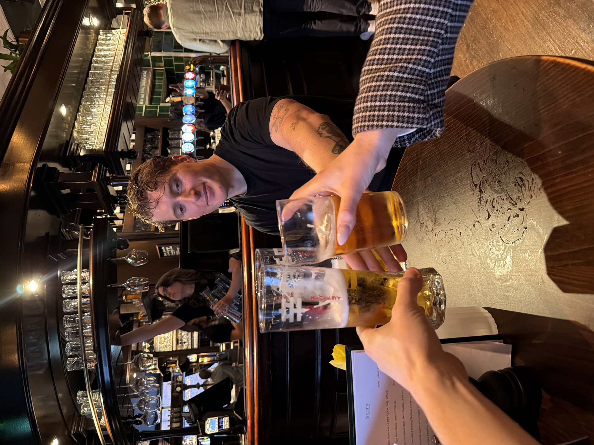 Here is a detailed description of the image:
**Scene:** The image captures a moment in what appears to be a traditional pub setting. A wooden bar is visible in the background, replete with an array of bottles and glasses neatly arranged.
**People:** There are two individuals in the foreground. One is a man facing the camera, wearing a black t-shirt, with visible tattoos on his arm. Another person is seen from the arm down, pouring beer into two glasses. This person is wearing a jacket with a black and white checkered pattern. Other patrons are visible in the background, albeit less distinctly.
**Objects:** Two glasses are being filled with beer, with the act of pouring being the central focus. A menu is positioned near the glasses, with the heading "WHITE" visible. Other notable objects include a black item (possibly a camera lens or part of a bag) and the wooden table on which the glasses are placed. The bar in the background features rows of bottles and an array of glasses above the bar.
**Actions and Interactions:** The primary action is the pouring of beer from one glass to another. There is a sense of camaraderie or celebration, evidenced by the act of toasting or sharing a drink. The man in the black t-shirt is looking towards the camera with a smile, suggesting engagement with the moment.
**Environment:** The overall ambiance suggests a cozy pub environment. This type of setting is common in London.