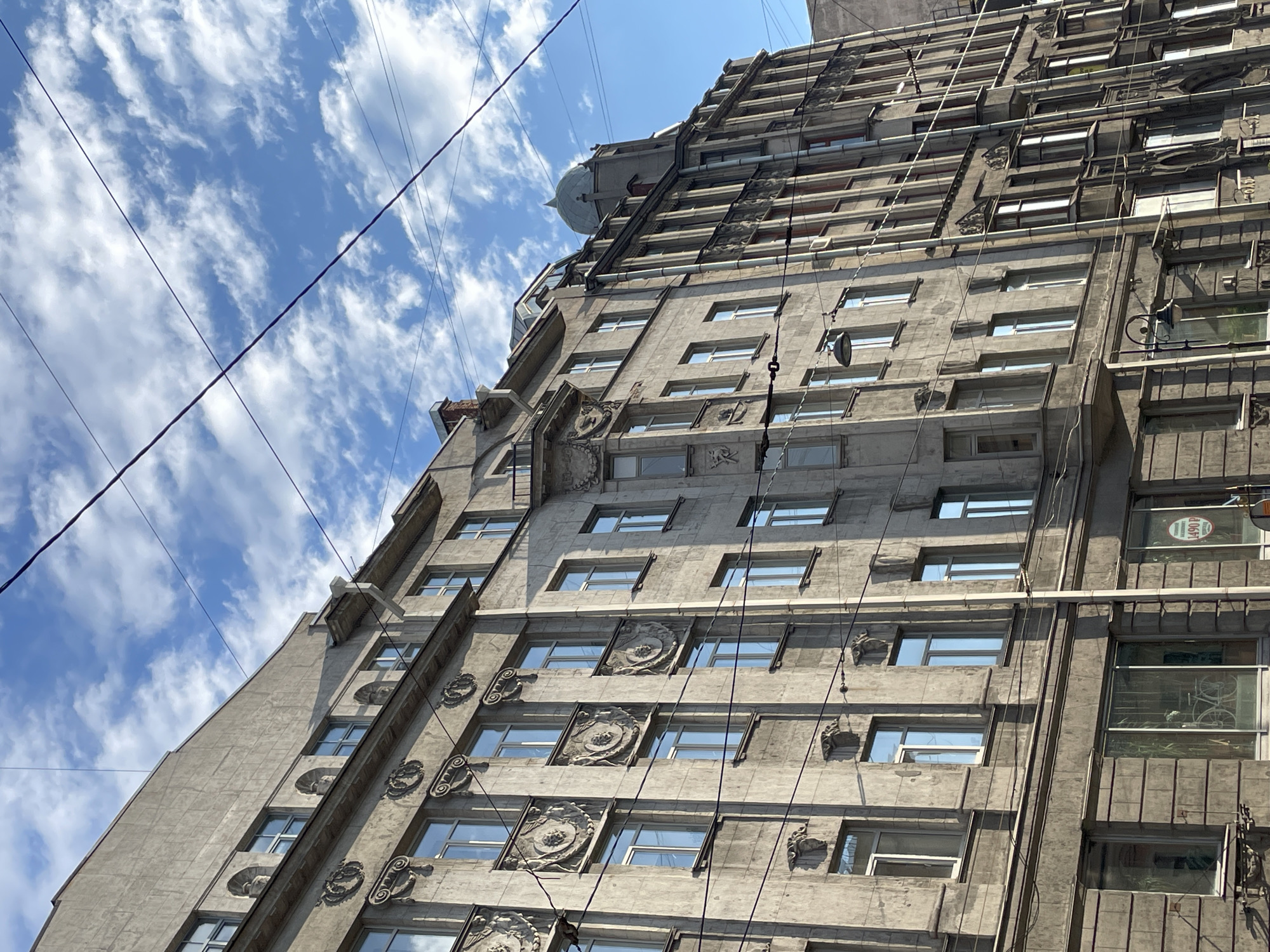 The image is a low-angle shot of a tall, gray building in Saint Petersburg, Russia. The building is adorned with intricate architectural details, including decorative friezes, medallions, and cornices. The facade features rows of rectangular windows reflecting the sky. Electrical wires crisscross the upper portion of the frame, contrasting with the ornate structure. The sky is blue with scattered white clouds. A small dome is visible on the building's roof.