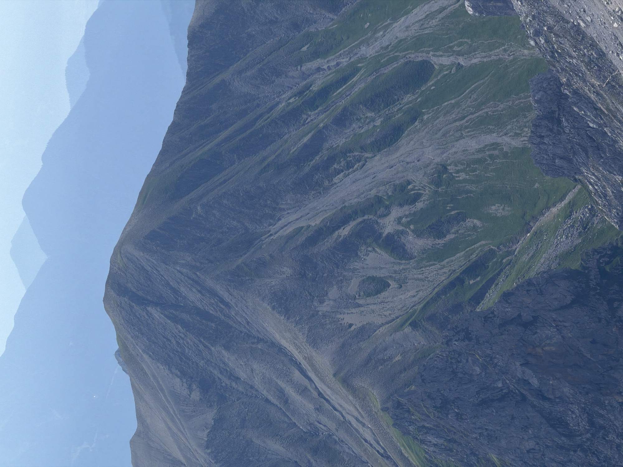 The image is a wide-angle, high-altitude shot of a mountain range. The foreground is dominated by the steep, rocky slopes of a large mountain. The slopes are a mixture of grey rock and patches of green vegetation. There are visible ravines and scree slopes on the mountainside. In the middle ground, the peak of another, less steep mountain is visible, also covered in vegetation and rock. The background consists of several layers of progressively paler, hazy blue mountains receding into the distance. The sky is a very pale blue, almost white. The scene appears to be a natural landscape, likely in a mountainous region, and is consistent with the topography of the area around Garmisch-Partenkirchen, Germany, known for its Alpine scenery. No people or man-made structures are discernible in the image. The lighting suggests it is daytime, with diffused sunlight.
