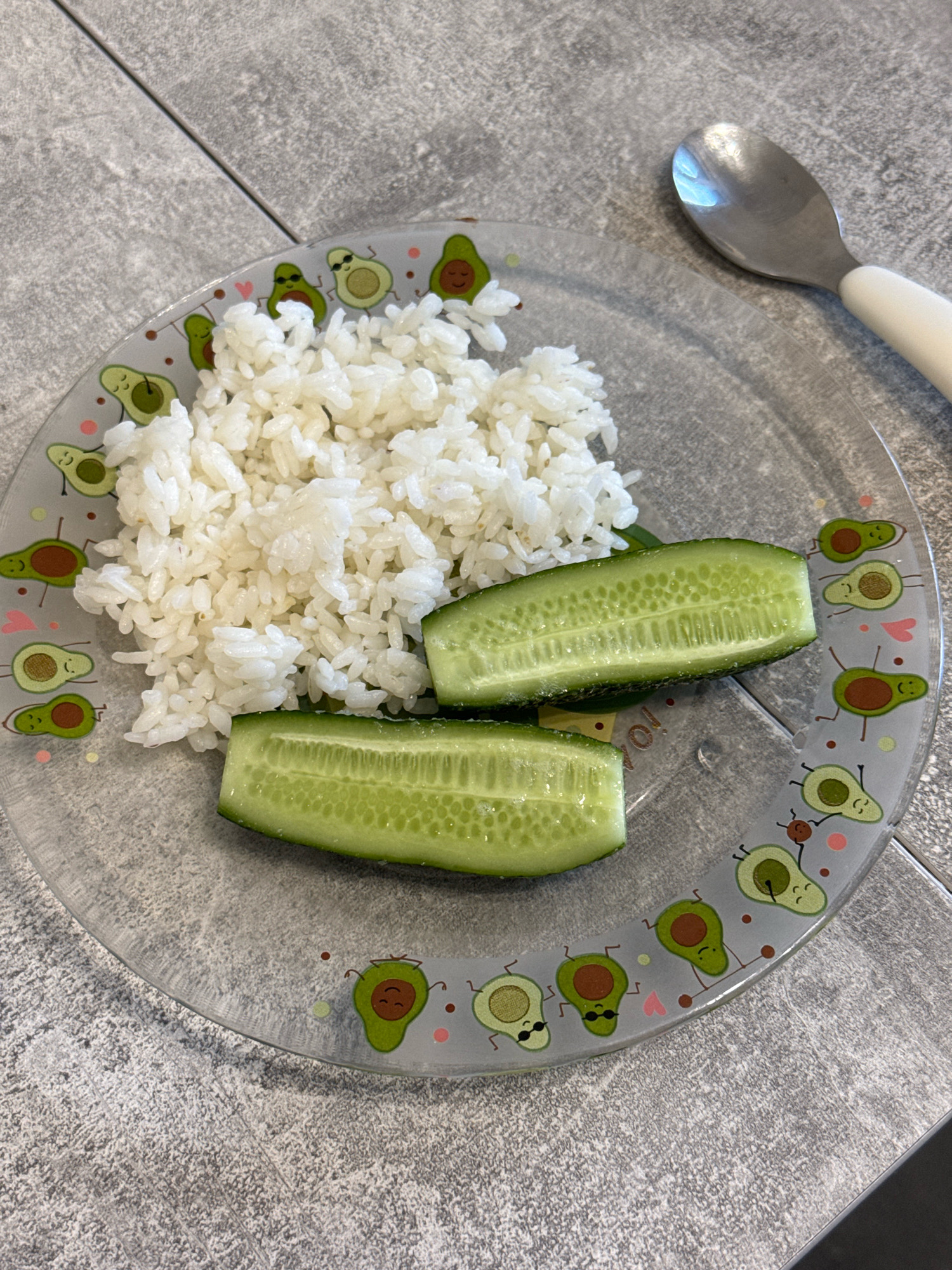 A clear glass plate decorated with cartoon avocados is filled with white rice and two slices of cucumber. A metal spoon with a white handle rests on the right side of the plate. The plate sits on a gray, textured surface, likely a table. The image does not contain any information about the city of Lyubertsy, Russia.
