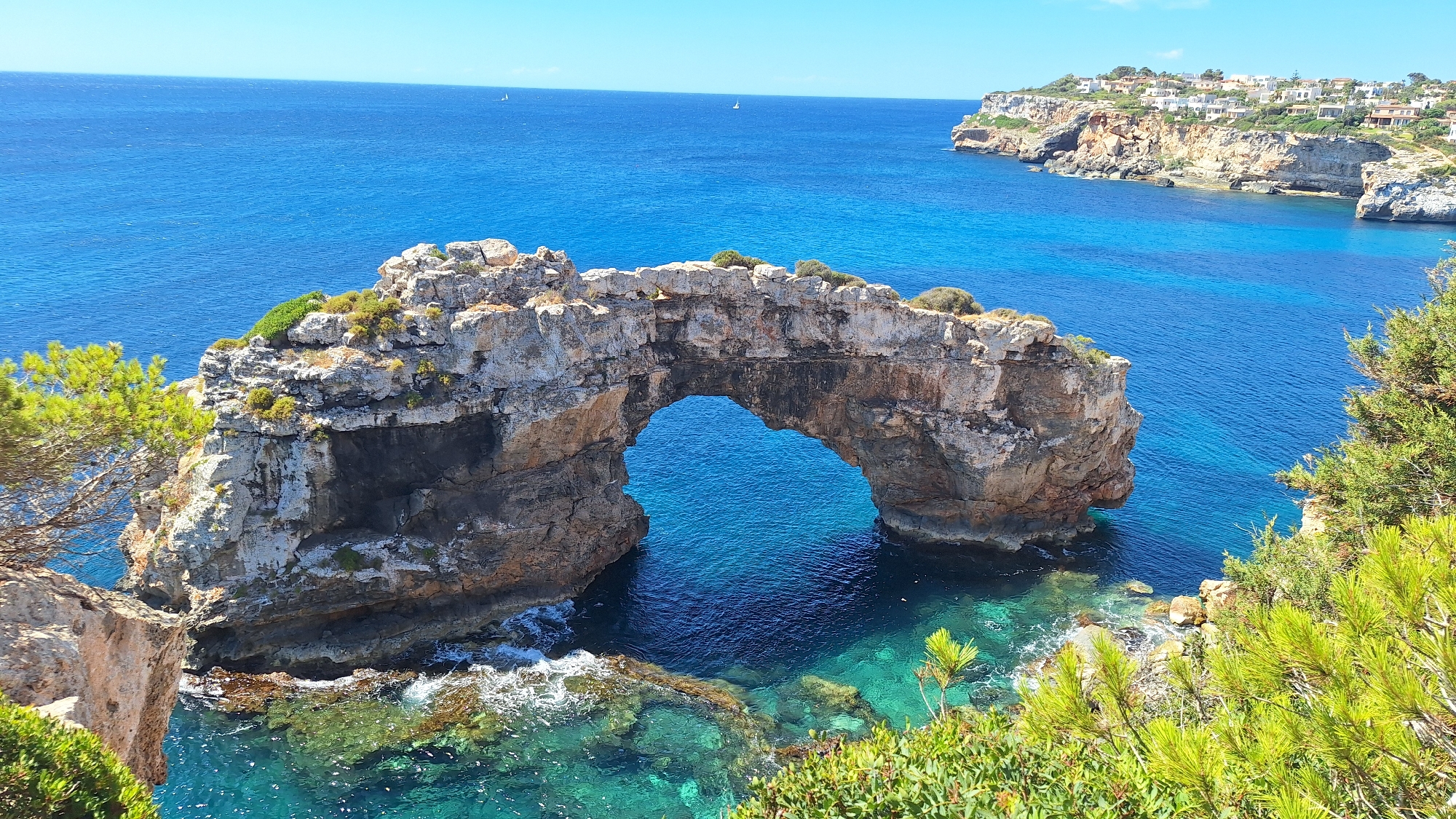 The image depicts a natural rock arch formation on a rocky coastline overlooking a bright blue ocean. The arch, composed of weathered stone, frames a section of the sea, revealing clear, turquoise water with visible rocky outcrops beneath the surface. The surrounding cliffs are rugged and covered with sparse vegetation, including green shrubs and pine trees. In the distance, the vast blue ocean stretches to the horizon, with two small sailboats visible. Along the upper right edge of the image, a cliff face is topped with buildings that appear to be houses, part of a coastal settlement. The sky is a clear, cloudless blue. The location is identified as being in the city of Santanyí, Spain, which is known for its coastal beauty. There are no people or animals visible in the scene. The overall environment is sunny and serene, emphasizing the natural beauty of the seascape and coastal landscape.