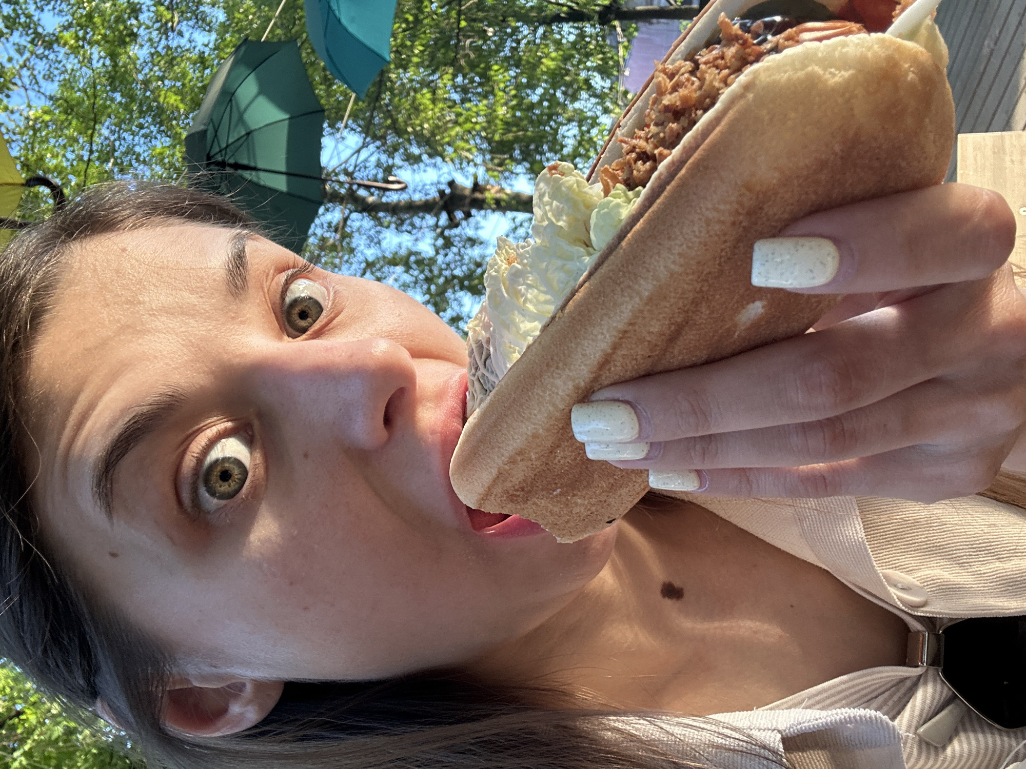 A woman is holding and eating a large sandwich. The sandwich appears to have a substantial amount of filling, including shredded meat and white topping such as sour cream. The woman’s eyes are wide, and she appears to be enjoying the food. The woman has a mole on her neck. The background shows trees and a few hanging umbrellas. The location is Izhevsk, Russia.