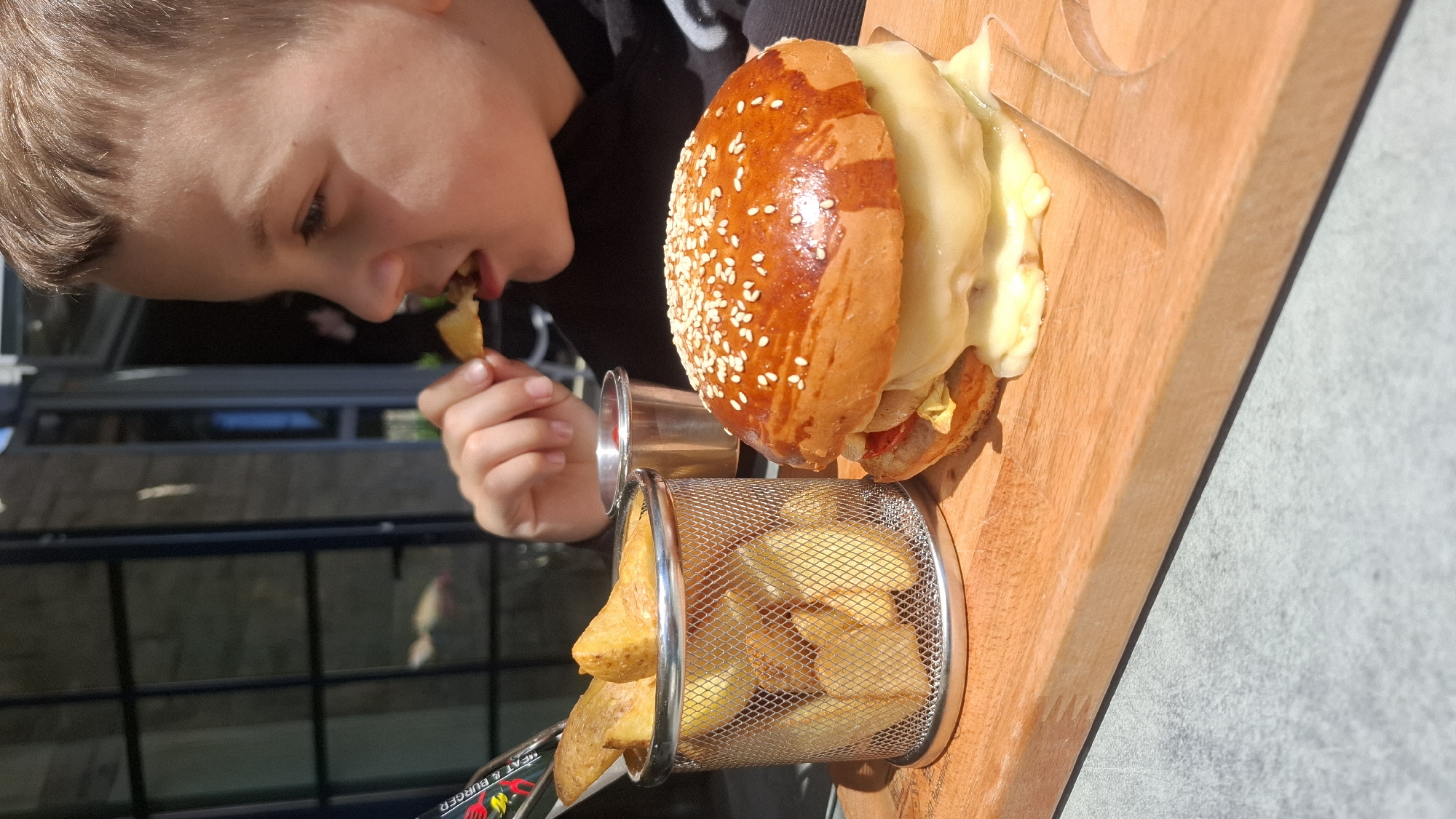 A young person is eating a fry from a wire basket placed on a wooden platter alongside a hamburger slathered in cheese. The hamburger has a sesame seed bun. The platter is resting on a gray surface outdoors, presumably in the city of Bečići, Montenegro. The individual is facing the food, and a building is visible in the background.