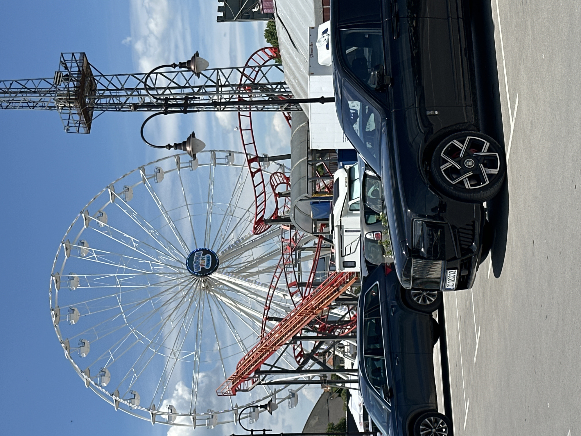 The image depicts a Ferris wheel and a roller coaster in Varna, Bulgaria, based on the "Varna Fun Park" logo on the Ferris wheel. The sky is blue with scattered clouds. A metal structure resembling a tower is also visible. In the foreground are parked cars, including a black SUV with a distinctive wheel design and a license plate that reads "CC 1200C2". The image appears to be taken from a parking lot adjacent to the amusement park. Street lamps are present along the side of the road facing the amusement park. The image has been rotated 90 degrees clockwise.
