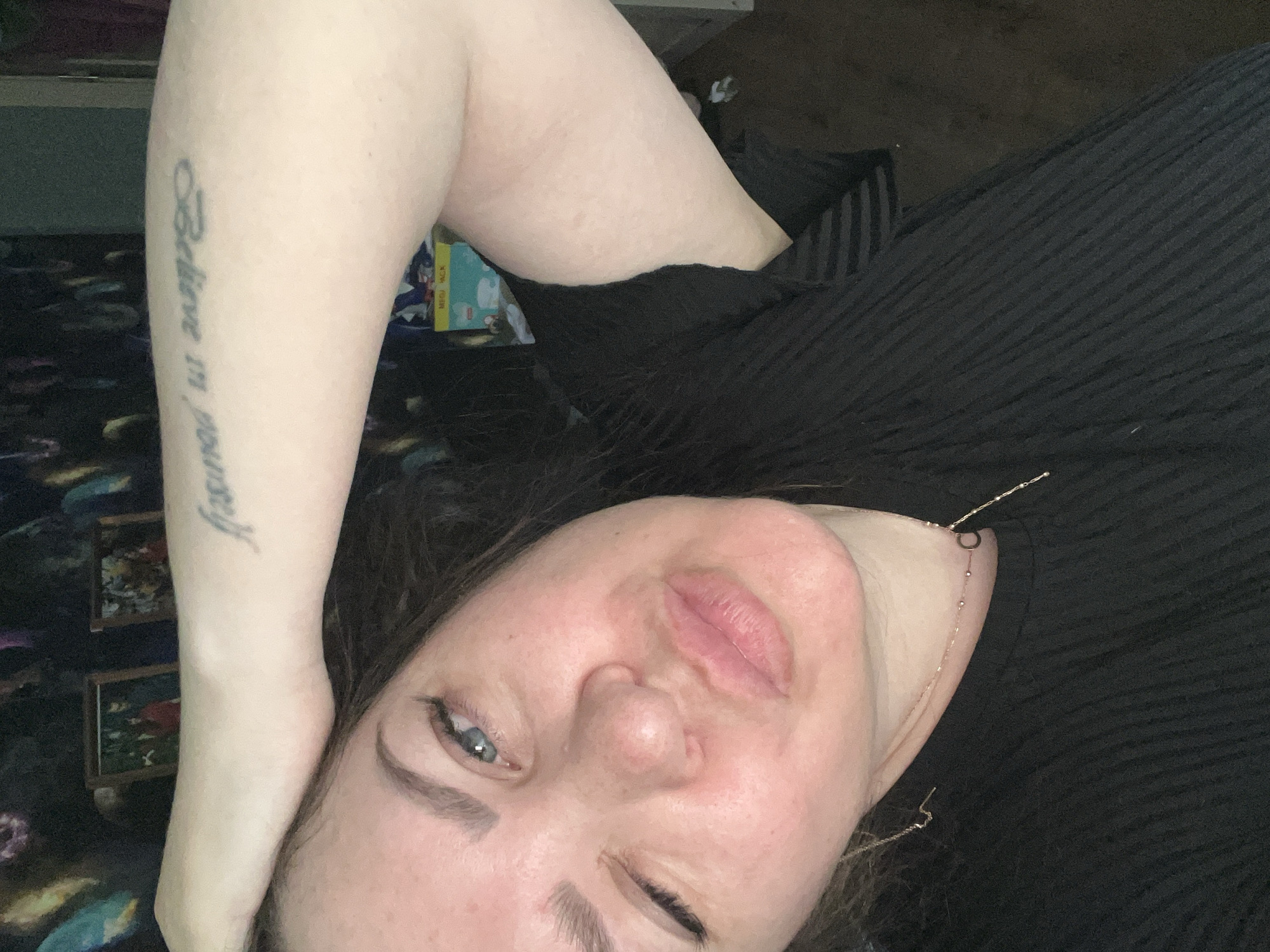 A person lies with their arm above their head. The person's arm has a tattoo that says "Believe in yourself." The person is wearing a black shirt and a gold chain. The person is lying on a black surface. There are framed pictures in the background.