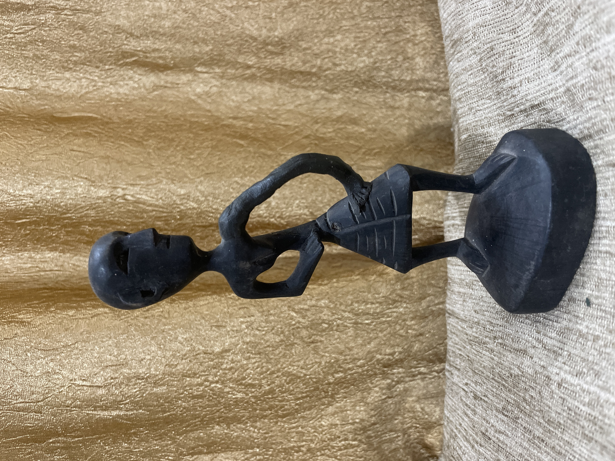 A dark, stylized wooden figurine of a person stands on a textured, beige surface in front of a shimmering gold fabric backdrop. The figurine has a slender body, elongated head with carved facial features, and arms bent at the elbows. It is wearing a conical skirt with horizontal lines. The figurine is positioned slightly to the right of the center of the image. The background is a wrinkled gold fabric with a crinkled texture that reflects light. The foreground shows a coarse woven beige material. No specific location in Bhagsu, India is identifiable.
