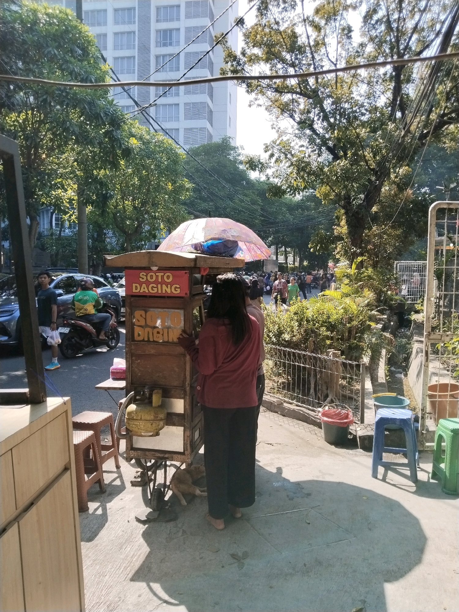 The image depicts a street scene in Surabaya, Indonesia, likely during the daytime, as indicated by the strong shadows and bright sunlight. In the foreground, a food cart selling "Soto Daging" (beef soup) is prominently displayed. The cart is made of wood and has a gas cylinder underneath. A woman in a maroon top and dark pants stands behind the cart, appearing to be engaged in serving or preparing food. A small dog is lying on the ground near the cart.
To the left of the food cart, a man in a dark t-shirt and white shorts stands near a parked motorcycle. The motorcycle has a rider wearing a green and yellow Grab uniform. In the background, there is a busy street with more people and vehicles, suggesting a commercial or residential area with moderate foot traffic. Trees line the street, providing shade, and a tall modern building is visible in the distance, characteristic of urban landscapes. Several plastic stools of various colors are placed along the sidewalk, some occupied and some empty. The overall environment appears to be an outdoor public space in a city.