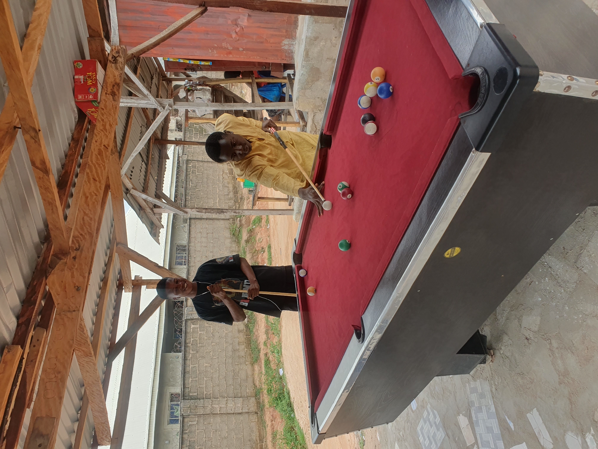 The image depicts an outdoor billiard area in Lokoja, Nigeria, characterized by a rustic, partially covered wooden structure. Two young African men are present. The man on the left, wearing a black t-shirt, stands behind a red-felted billiard table, holding a pool cue. The man on the right, dressed in a yellow robe, is actively playing, striking a billiard ball with his cue. The table has several billiard balls scattered on its surface, including a white cue ball, solid colored balls, and striped balls. The environment appears to be an informal gathering space, possibly a recreational area or an establishment in Lokoja. The background shows a brick wall and some vegetation, suggesting an urban or semi-urban setting. The overall scene captures a moment of leisure and friendly competition.