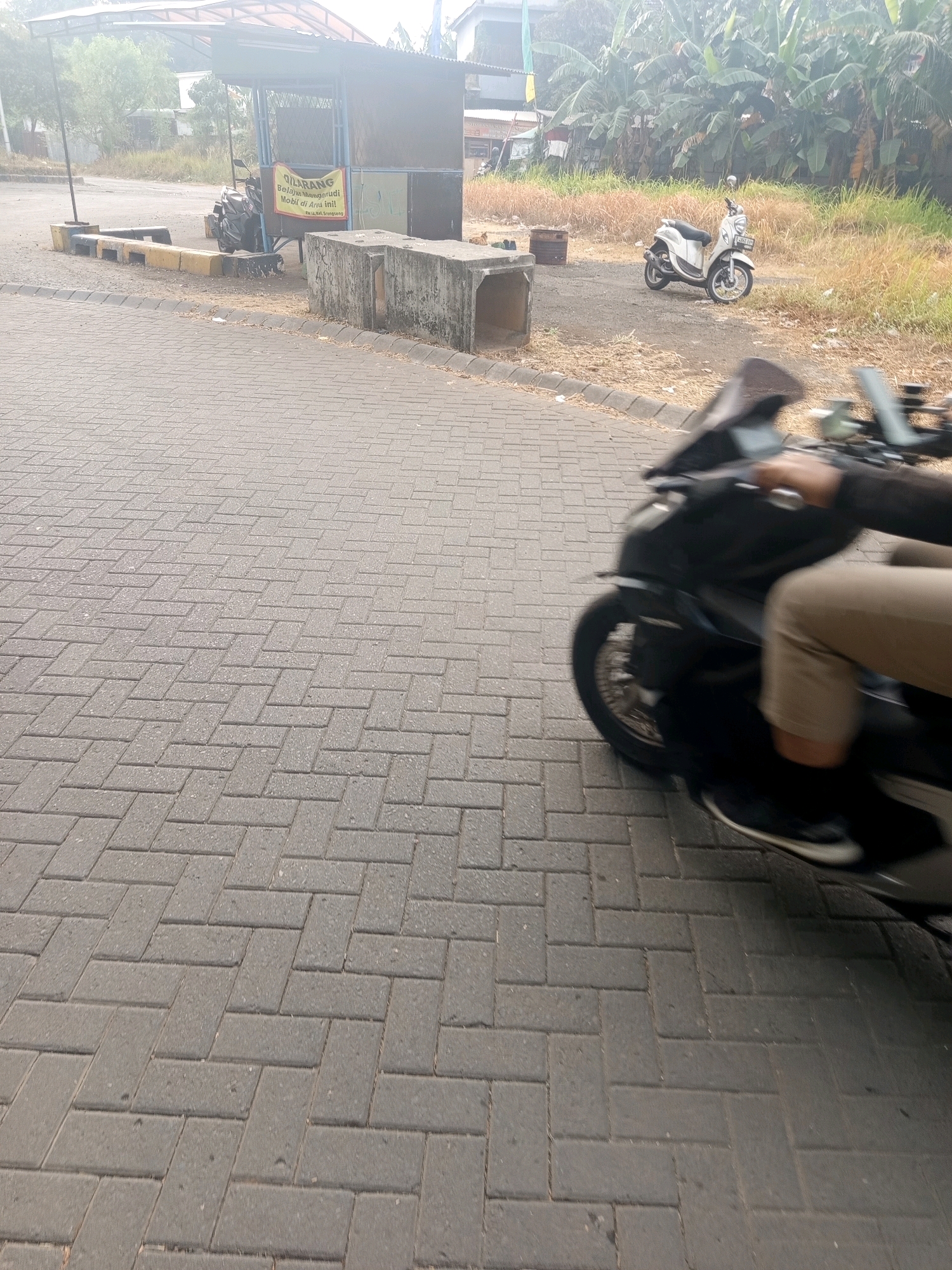 The image captures a street scene, likely in West Jakarta, Indonesia, characterized by its paved brick walkway and a backdrop of vegetation. In the foreground, a person is riding a dark-colored motorcycle, their legs and torso visible, suggesting motion. The motorcycle's wheel is blurred, indicating movement. Further in the background, a white scooter is parked to the right of a small shelter with a yellow sign. A darker motorcycle is parked to the left of the shelter. The environment appears to be a somewhat rural or suburban area, with abundant greenery and some structures suggesting urban development. The overall lighting suggests it could be daytime, possibly in the morning or late afternoon given the slight haze.