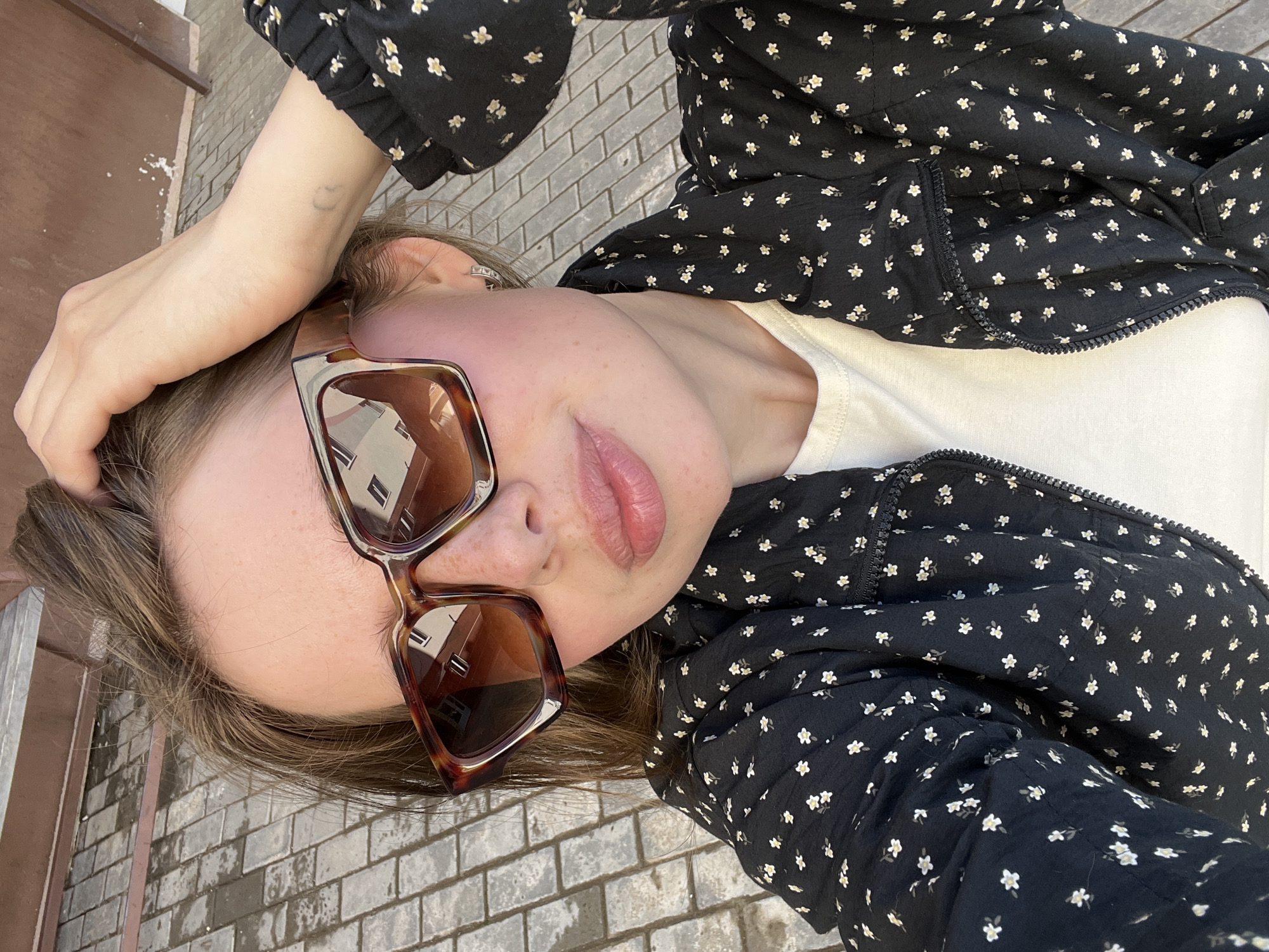 A young woman with light brown hair, wearing large, square, tortoiseshell sunglasses and a black jacket with a white floral pattern over a white t-shirt, poses for a selfie. She is outdoors, with her right hand in her hair. The background shows grey brick paving stones and a blurred brown wall. The image appears to have been taken during daylight. The reflection in her sunglasses shows buildings. The tattoo on her left forearm is visible and appears to be a faint outline of a heart and possibly another small symbol. The overall scene suggests a casual outdoor setting in a city.