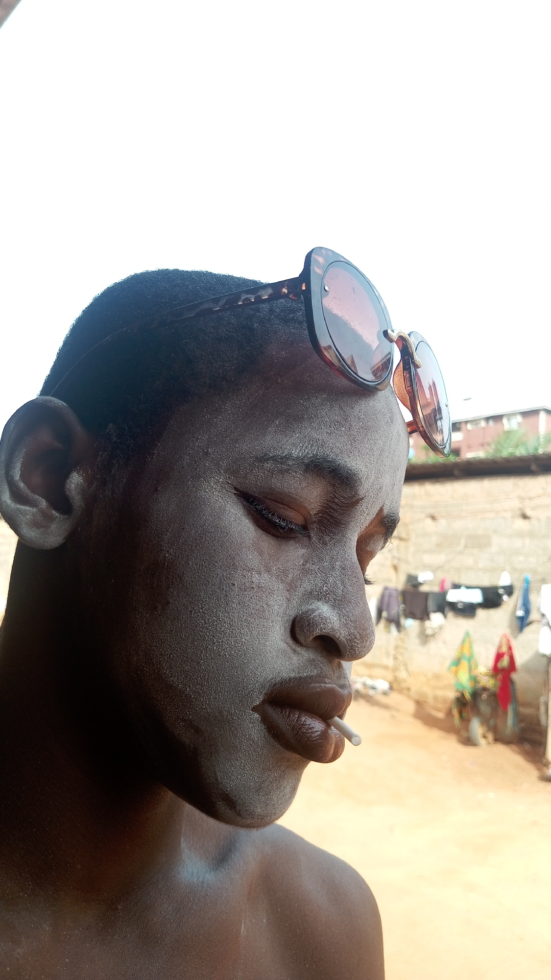A close-up, low-angle, profile view shows a dark-skinned person with sunglasses perched on their head and a thin, white object in their mouth. The person's face appears to be covered in a light-colored powder. A blurred background reveals a sunlit outdoor space with a wall, laundry hanging to dry, and two brightly colored items that could be umbrellas or decorations. A building is visible in the far background. The location is listed as the city of Unknown, Unknown.