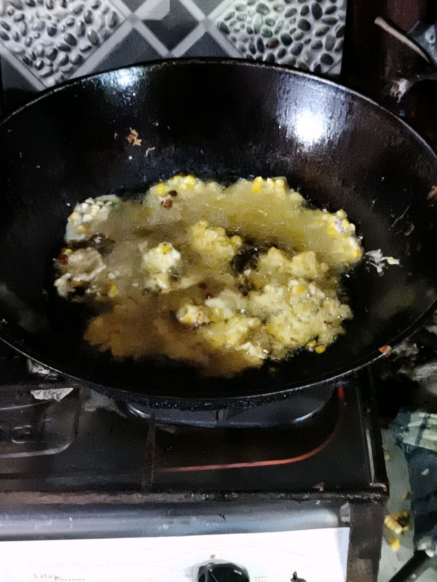 The image shows a close-up of a black wok filled with food being deep-fried in hot oil. The food appears to be a mixture of corn and batter, with some reddish specks which could be chilies or other spices. The wok is placed on a black gas stove. In the background, a tiled wall with a pattern of black and grey oval shapes is visible. The lighting suggests the photograph was taken indoors. The scene is likely a kitchen in Betelan, Indonesia, given the culinary context and common household items depicted.