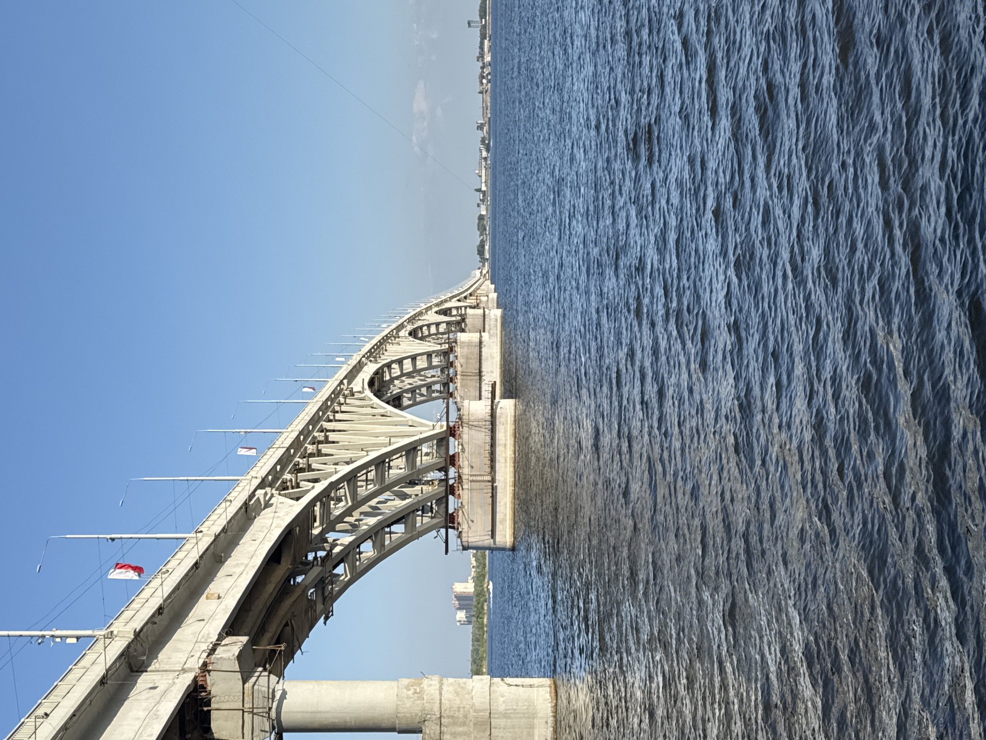 A long, concrete and steel arched bridge spans a wide, calm river under a clear blue sky. The bridge is supported by large concrete pillars and has numerous lights running along its edge. Two flags, one red and white, and another predominantly white, are visible on the bridge. In the distance, across the river, a city skyline with buildings and trees can be seen. The water in the foreground is rippled and dark blue. The scene appears to be the Saratov Bridge in Saratov, Russia.