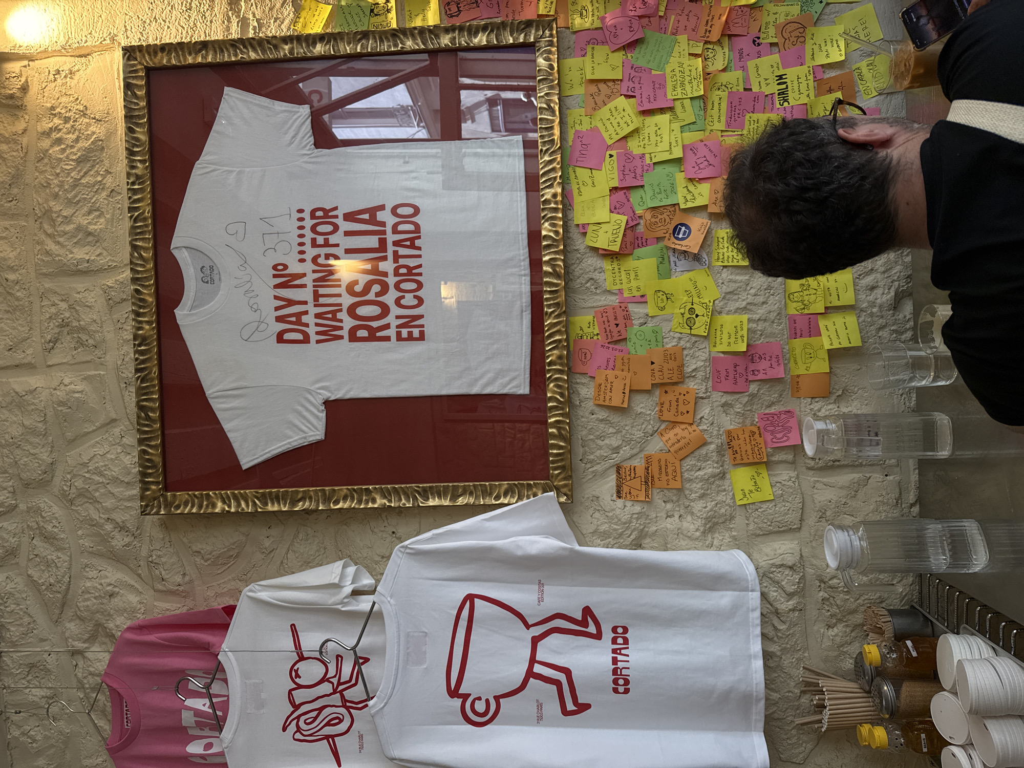 A medium, eye-level, indoor shot shows an off-white stone wall covered with sticky notes, t-shirts, water pitchers, and a male figure.
The wall is covered with multi-colored sticky notes bearing handwritten messages. A framed white t-shirt hangs above a set of three t-shirts on hangers. The framed t-shirt is white with red lettering that reads "DAY NO 371 WAITING FOR ROSALIA EN CORTADO." There is a silver autograph above the lettering. The lower t-shirts are on hangers and display the text "Cortado." To the right, glass water pitchers stand on a shelf next to a metal bin holding condiments, wooden stir sticks, and cups.
To the right, the back of a man's head is visible, and he is wearing black-rimmed glasses, a white strap (possibly of a bag), and a black jacket. His dark, short, curly hair is visible. He appears to be looking at the wall.
The location is Paris, France. The t-shirts appear to reference a Cortado coffee shop.