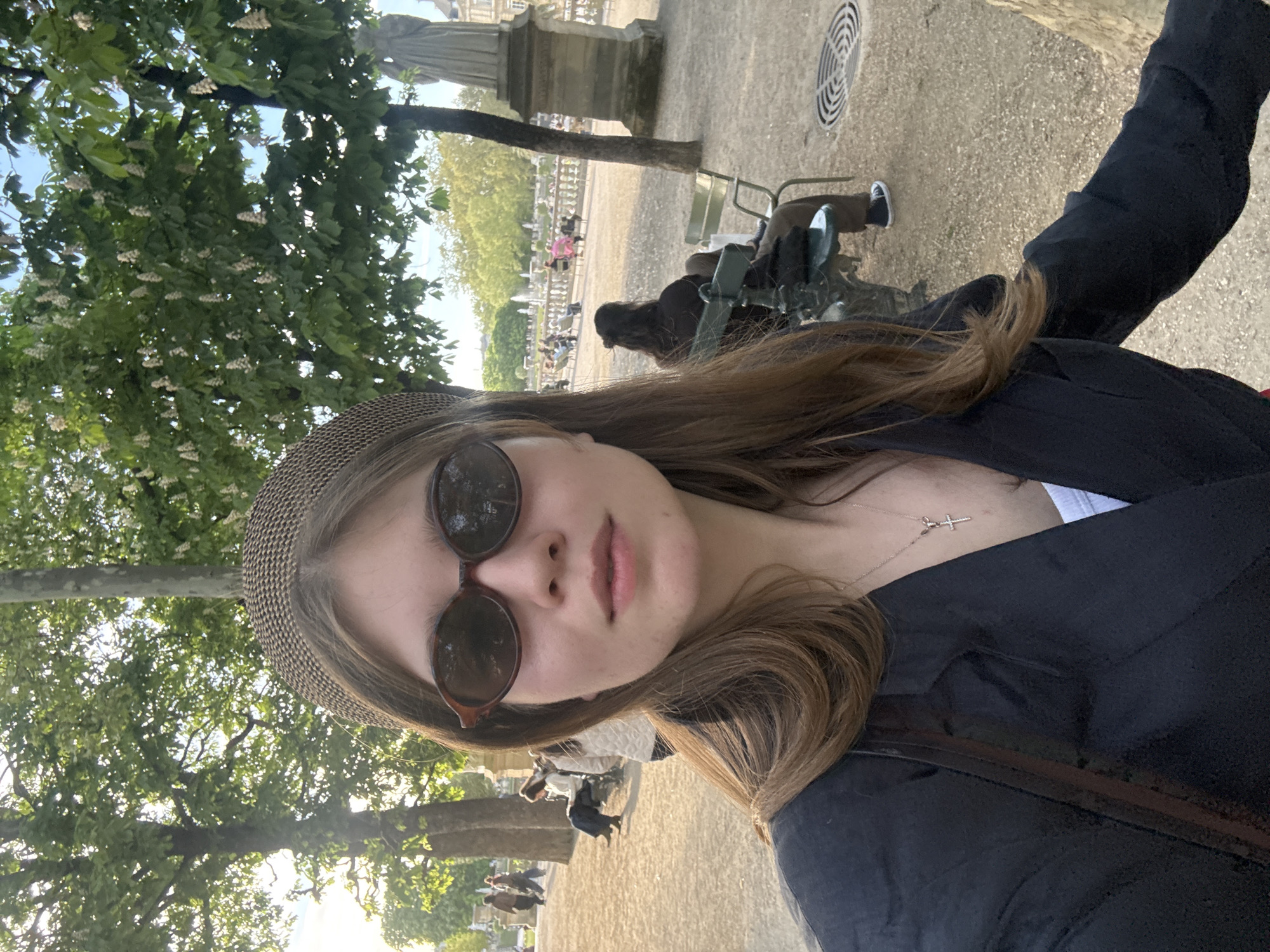A selfie shows a woman wearing a hat and sunglasses, with a crucifix necklace visible. In the background is the Jardin du Luxembourg, a park in Paris, France. People are seen sitting on park chairs, and trees and a statue are visible. The lighting suggests it is daytime.