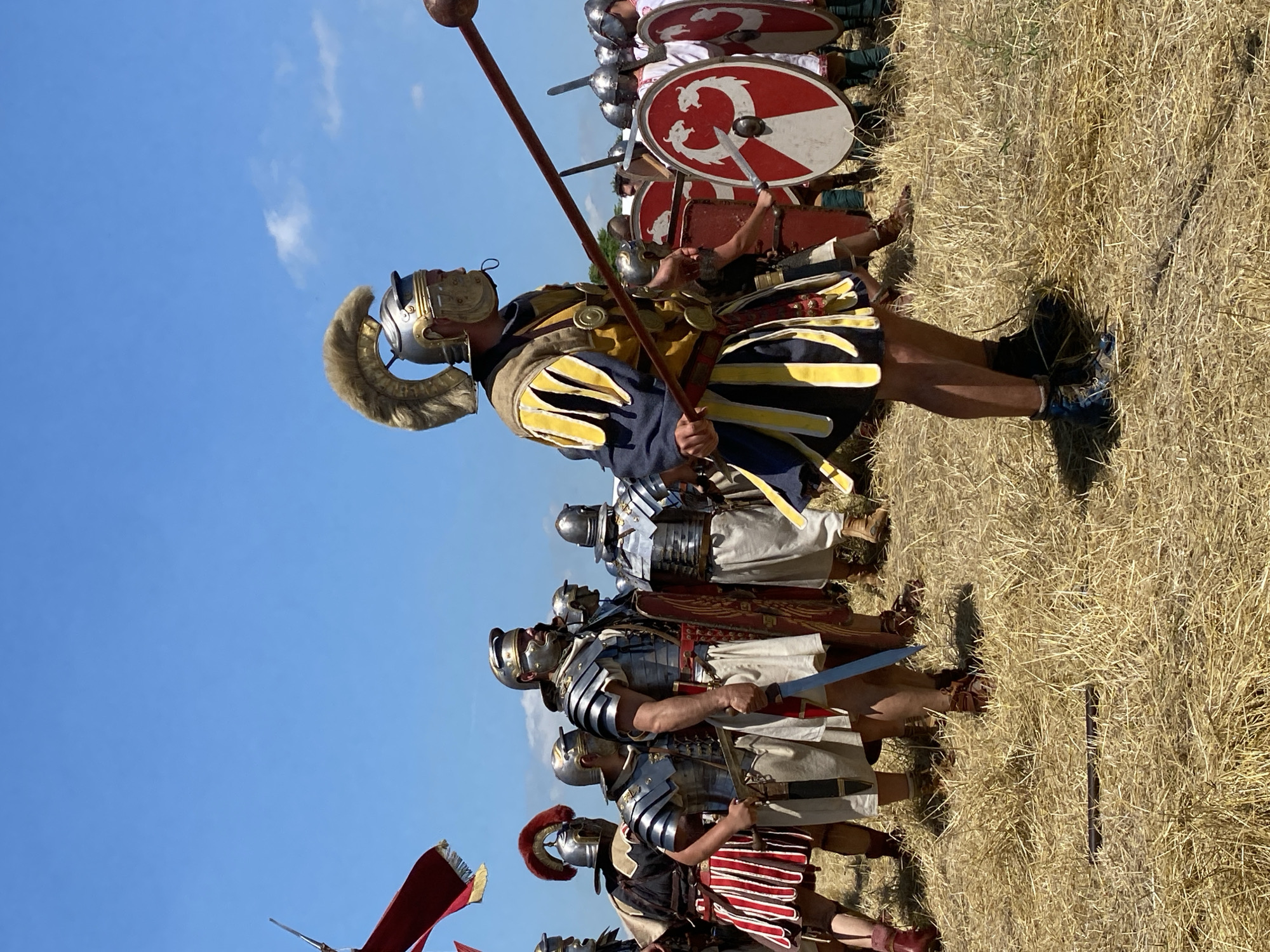 The image depicts a historical reenactment featuring individuals dressed as Roman soldiers in a field under a clear blue sky, located in Gigen, Bulgaria. In the immediate foreground, slightly right of center, a male figure is presented in profile, facing left. He wears a metal helmet with an elaborate, light-brown crest, a dark blue tunic featuring yellow and black vertical striped pteruges, and prominent shoulder armor, while holding a long spear or pilum diagonally across his body with a spherical counterweight. To the left of this primary figure, a group of approximately ten to fifteen other male figures are arranged in formation. Several wear segmented plate armor (lorica segmentata), metal helmets, and light-colored tunics, holding short swords (gladii). One figure towards the midground left wears a red-plumed helmet and a red and white striped skirt. Towards the midground and background right, additional figures are visible, similarly armored, with some carrying round shields (parmae) that display a red and white design featuring two intertwined white serpentine or dragon-like creatures. The ground beneath the individuals is covered with dry, light-brown straw or hay. The sky above is largely clear blue with minimal, wispy white cloud cover, indicating bright, sunny conditions. A portion of a red banner or flag is visible in the upper left corner of the frame.