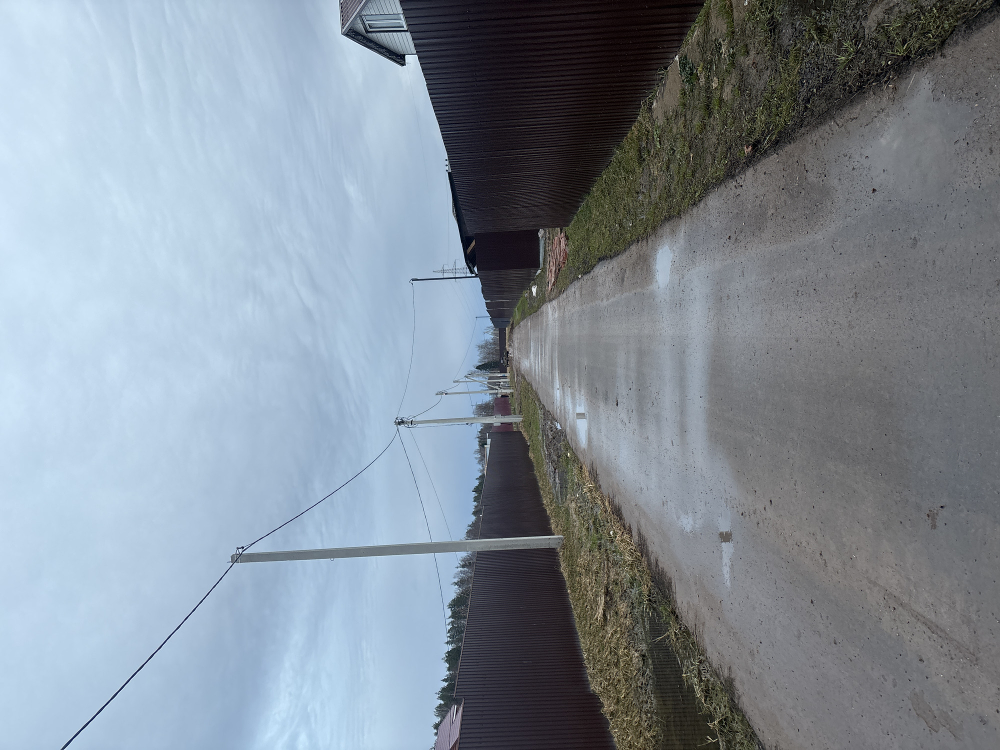 The image shows a wet, paved road in Раздолье, Russia, bordered by brown corrugated metal fences on both sides. Patches of grass and weeds grow along the edges of the road. Utility poles line one side, with wires stretching across the sky. The sky is overcast and gray. A corner of a building is visible in the upper left of the frame. No people or animals are visible.