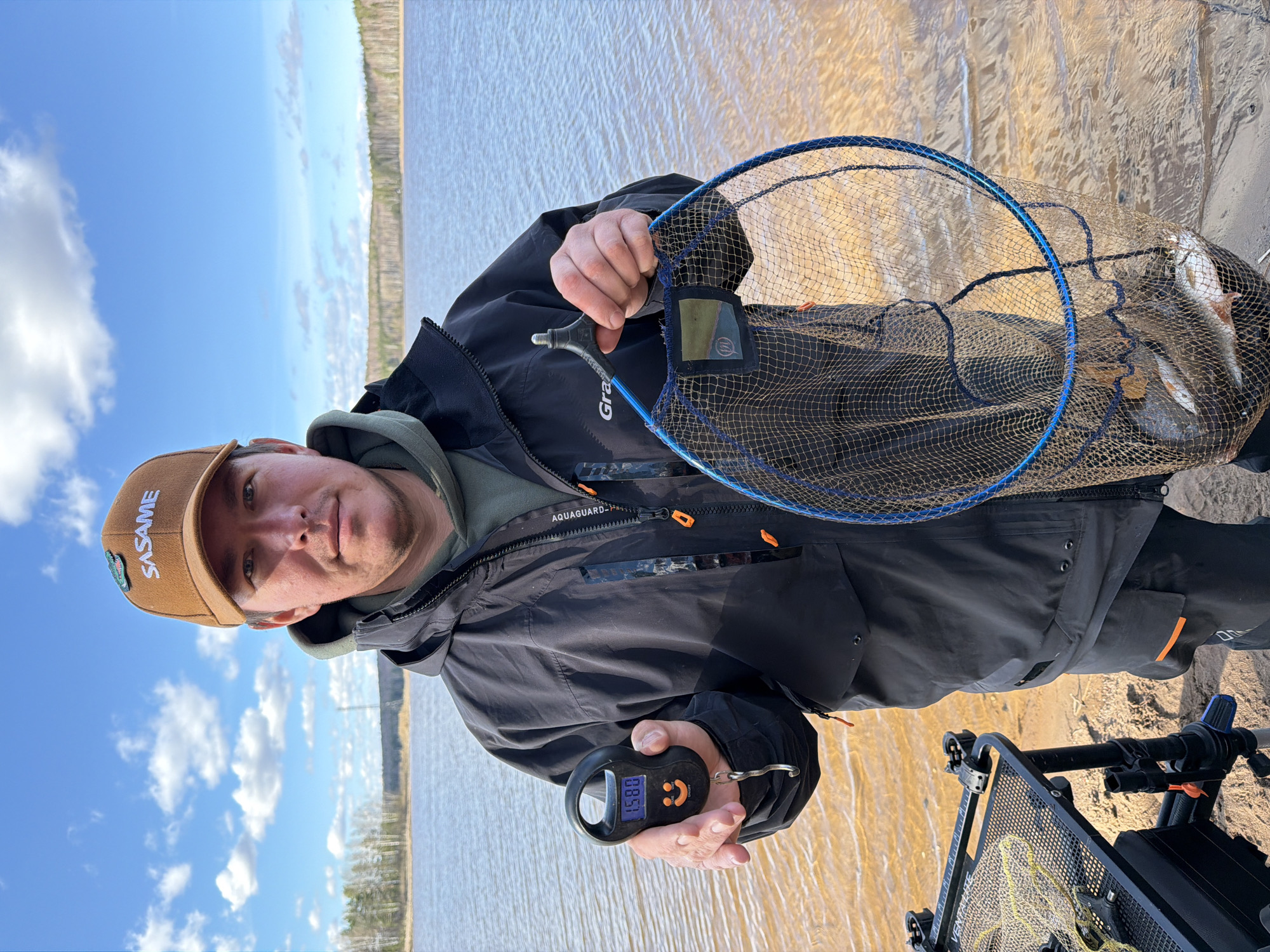 A man is standing near a body of water, possibly the city of Chaikovsky, Russia, based on the "SASAME" inscription on his hat. He is wearing a brown hat, a black jacket, and a green hoodie. In his left hand, he is holding a digital scale with a reading of "0.850". He is also holding a blue fishing net with several fish inside. The background shows water, sky, and trees. Near the bottom of the image is a black platform stand.
