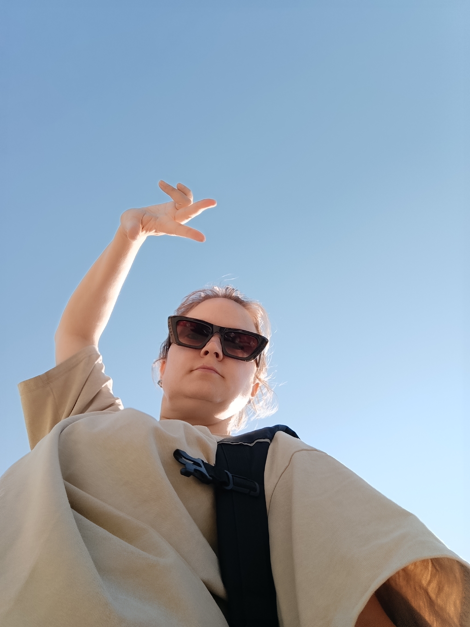 A low-angle, outdoor, full-body shot captures a person from the chest up, against a clear blue sky, suggesting a daytime setting possibly in the city of Chaikovsky, Russia. The individual, seen from below, is wearing sunglasses and a light-colored, loose-fitting t-shirt over a dark backpack with a black buckle visible on the t-shirt. Their left arm is raised with their hand open, fingers extended, in a gesture towards the sky. The light suggests the sun is behind the viewer, illuminating the person's face and arm from the front. The background is a uniform, cloudless sky.