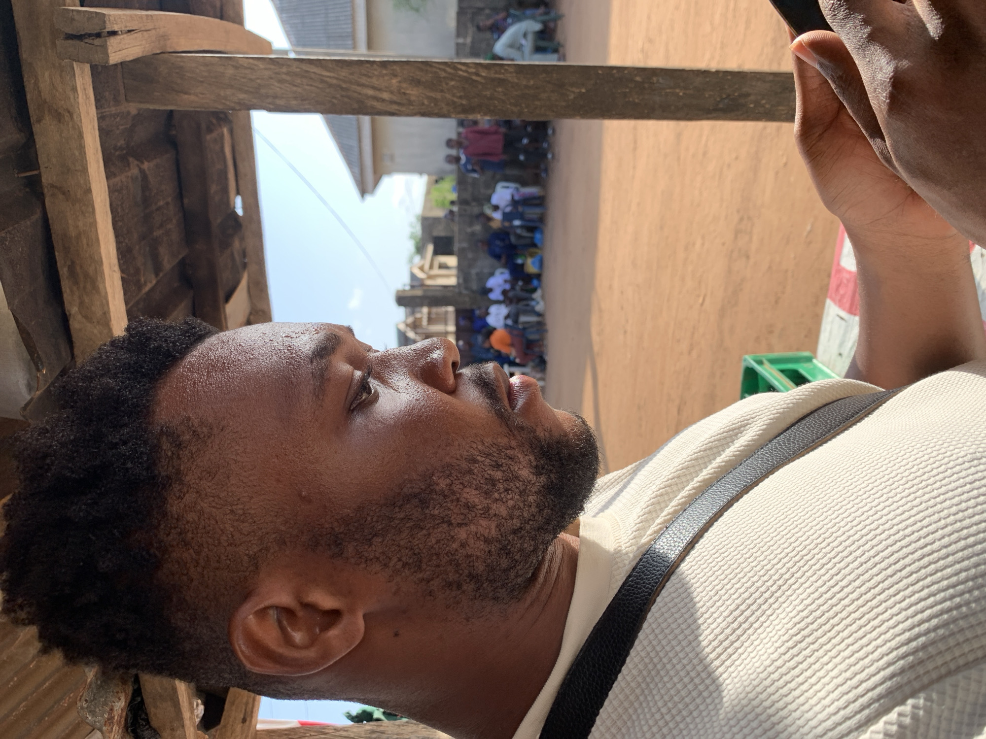 A man is shown in a medium close-up shot. He has dark hair and skin, a beard, and is wearing a light-colored shirt with a black strap across his chest. He's looking up and to the right. In the background, partially obscured by wooden beams, is a group of people gathered in what appears to be an outdoor area, possibly a dirt road or courtyard. Buildings can be seen in the distance. The picture's composition suggests the photo was taken from a low angle, possibly with the man holding a phone or camera above him to capture the scene. The setting is speculated to be in Ikorodu, Nigeria, based on details and visual cues from the image.