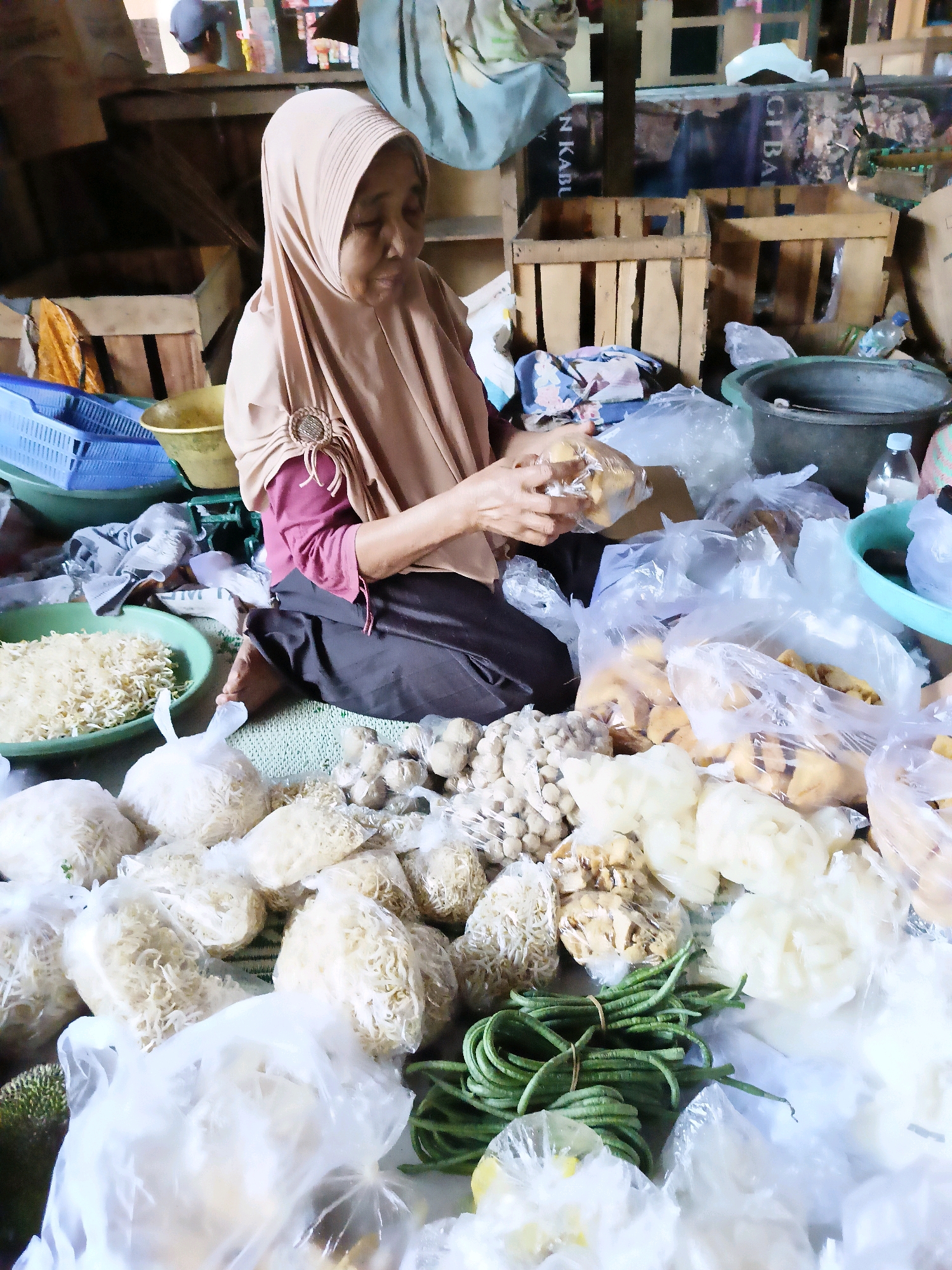 A woman wearing a hijab is seen in a market stall in Manisrenggo, Indonesia. She is sitting on a mat surrounded by various food items. The items are mostly packaged in plastic bags and include what appears to be dried noodles, some sort of processed food or snacks, and a bundle of green beans. In the background, there are wooden crates, baskets, and other market stalls. The woman is holding and examining some food items in her hands. The overall scene depicts a typical market environment with a vendor preparing or organizing her goods.