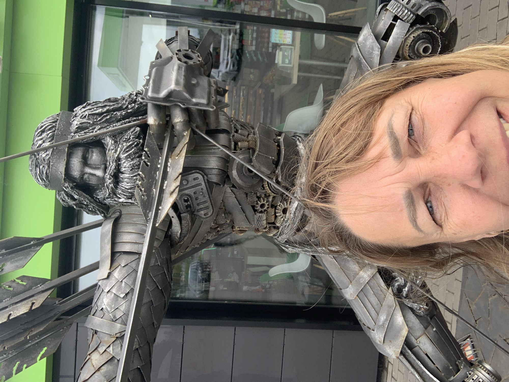 A woman with shoulder-length blonde hair takes a selfie in front of a large metal sculpture of an archer, which is located outside a building with a green facade and large windows, reflecting cars and tables with chairs. The sculpture is made of salvaged metal parts and depicts an archer with a bow drawn and arrows nocked. The woman's face, from the nose up, fills the foreground of the image. The location appears to be a roadside service area or a small shop.