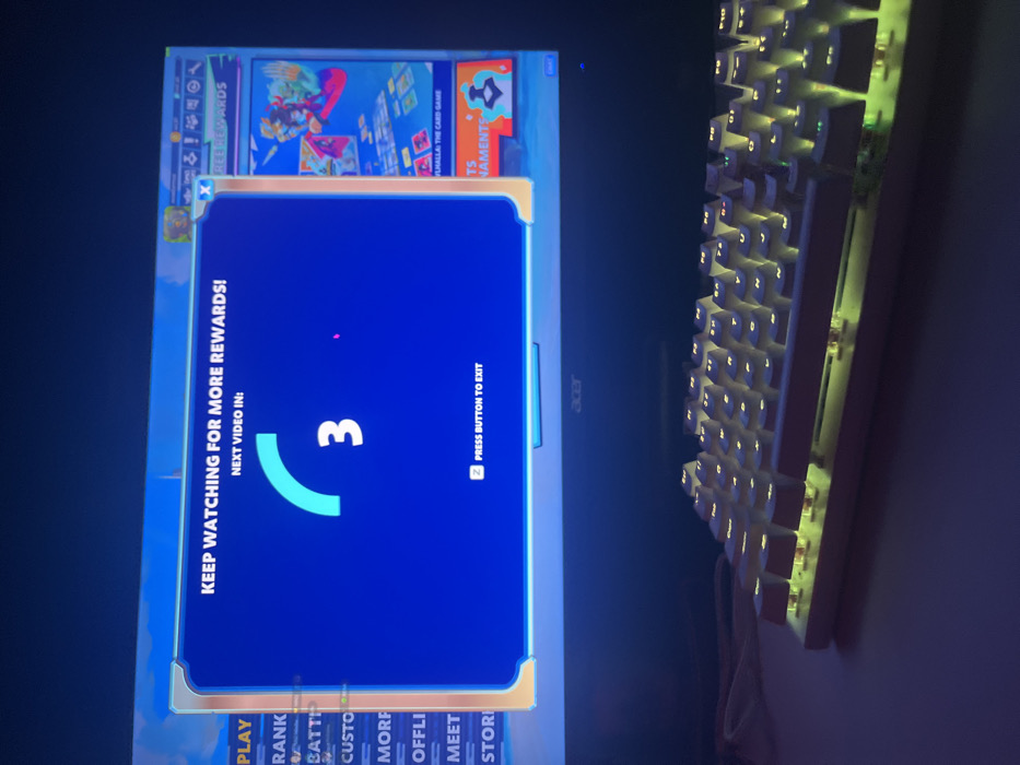 The image depicts a gaming setup located in Donchevo, Bulgaria. An Acer brand computer monitor, partially visible, displays a video game interface. A prominent blue rectangular overlay occupies the central screen area, featuring white text "KEEP WATCHING FOR MORE REWARDS!" and "NEXT VIDEO IN:", accompanied by a light blue circular progress bar indicating a countdown of "3." Below the countdown, the instruction "Z PRESS BUTTON TO EXIT" is visible. Portions of the underlying game interface are visible on the left and right, including menu items such as "PLAY" and "RANK," and a section labeled "FREE REWARDS" with artwork for "BRAWLHALLA: THE CARD GAME." Positioned on a flat, dark surface directly in front of the monitor is a white, compact mechanical keyboard. Its keys are illuminated with a yellowish-green backlight. The ambient lighting in the environment is low, with the monitor and keyboard providing the primary illumination. No individuals or active interactions are discernible within the frame.