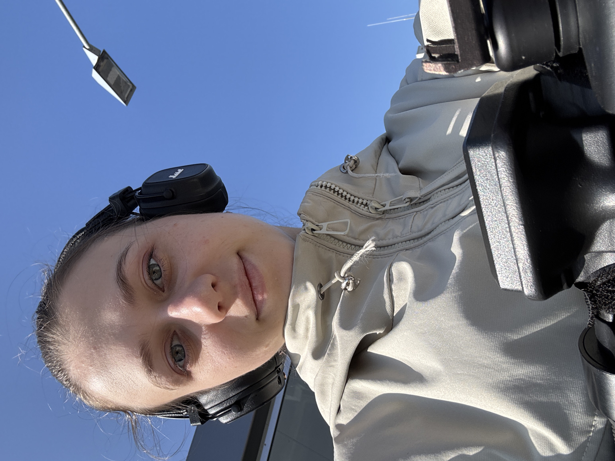 A young woman with blue eyes and blonde hair pulled back is taking a selfie outdoors. She is wearing black headphones and a light-colored jacket. The background is a clear blue sky and a street lamp. The scene is likely in Miltenberg, Germany, given the context of the image.