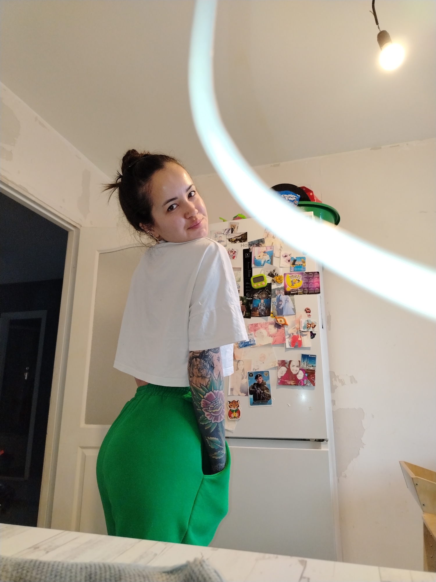 A woman stands in what appears to be a kitchen, facing away from the camera but looking back over her shoulder. She has dark hair pulled into a bun and is wearing a white, cropped t-shirt and bright green sweatpants. The woman has a detailed tattoo sleeve on her left arm.
Behind her is a white refrigerator covered with various magnets, photographs, and small items. The walls are white, with signs of repair work in progress, particularly towards the ceiling. A doorway to the left of the woman reveals a dark interior space. There's a light fixture visible in the upper right corner of the frame. A light colored countertop is visible in the foreground.
The image does not provide any indicators as to the location in the city of Noviy, Russia.